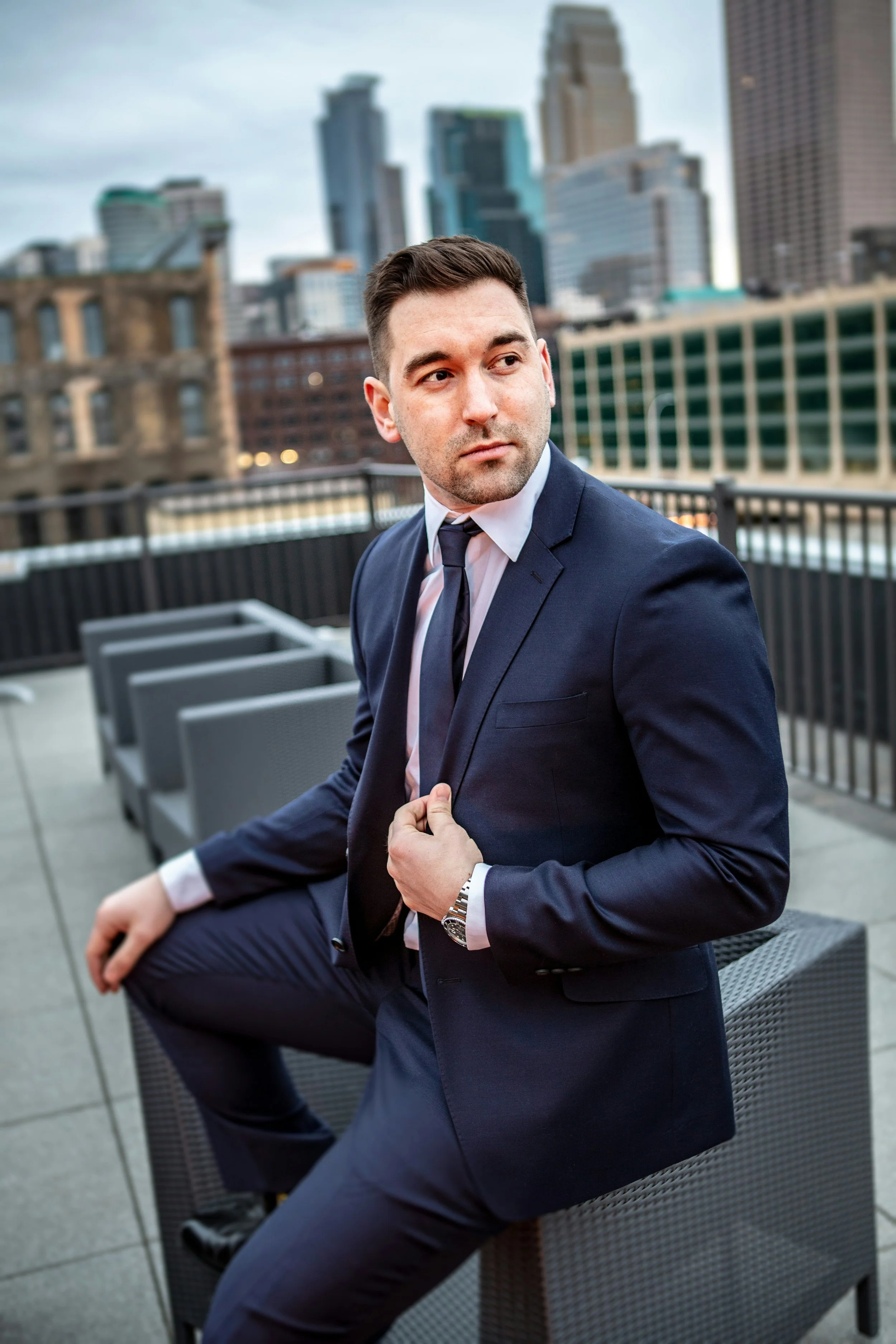 A man in a dark navy suit, white shirt, and dark tie sitting on an outdoor patio with city skyscrapers in the background. He is adjusting his suit jacket and looking to the side, with a wristwatch visible on his left wrist.