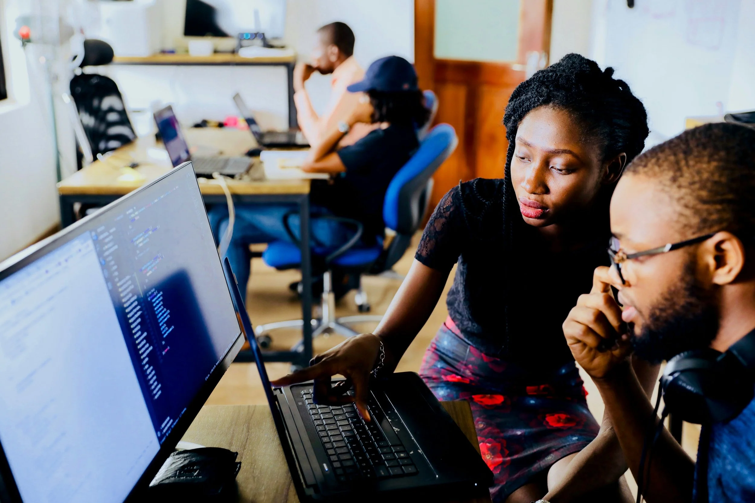 Three people working on laptops in an office or classroom. Two are in the foreground, focused on the screen, and one is in the background with a hand on his face, all engaging with their computers.