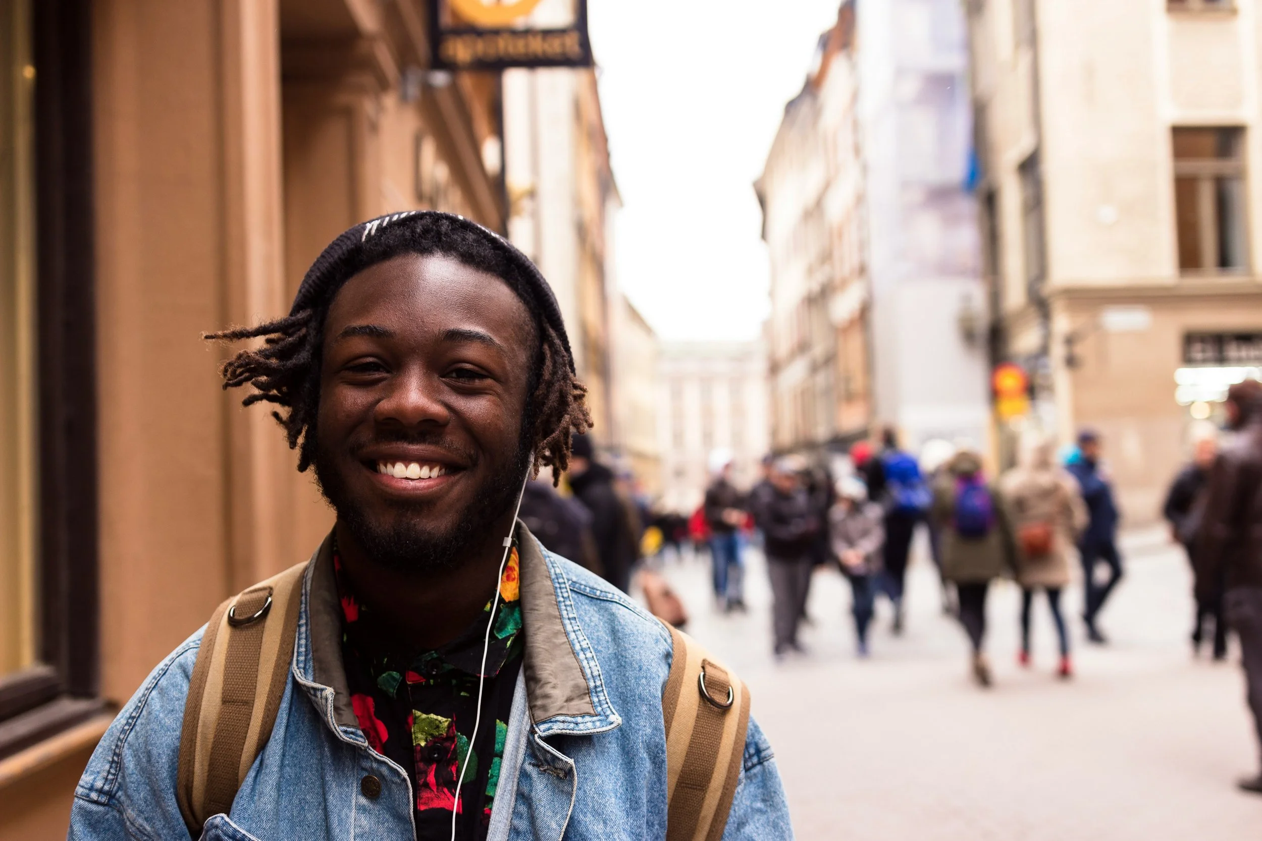 Smiling young man with dreadlocks, denim jacket, backpack, and earphones on a busy city street with blurred pedestrians and buildings.