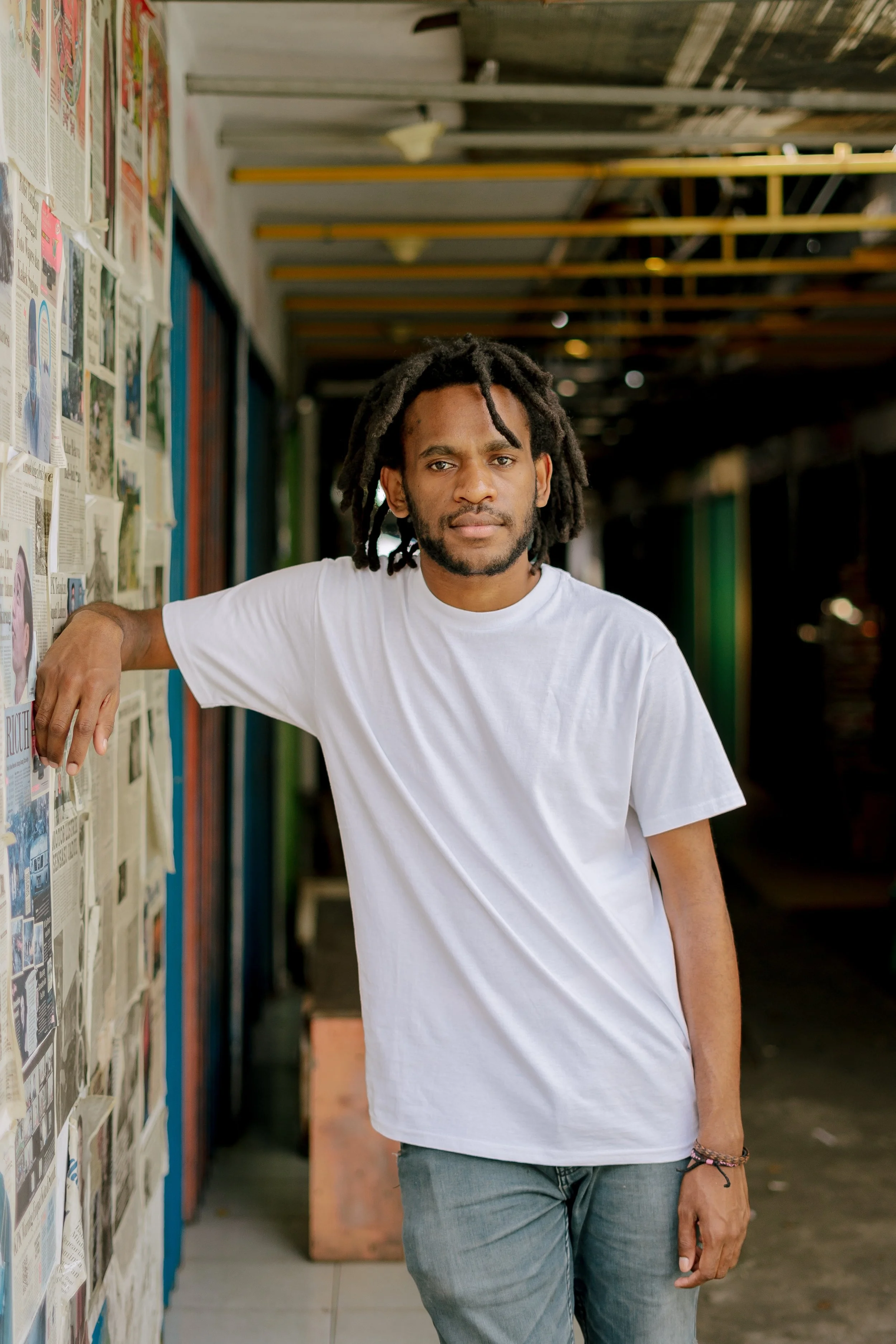 A young man with dreadlocks and a beard, wearing a white t-shirt and jeans, leaning against a wall covered with newspapers or magazines in an indoor or semi-outdoor setting.