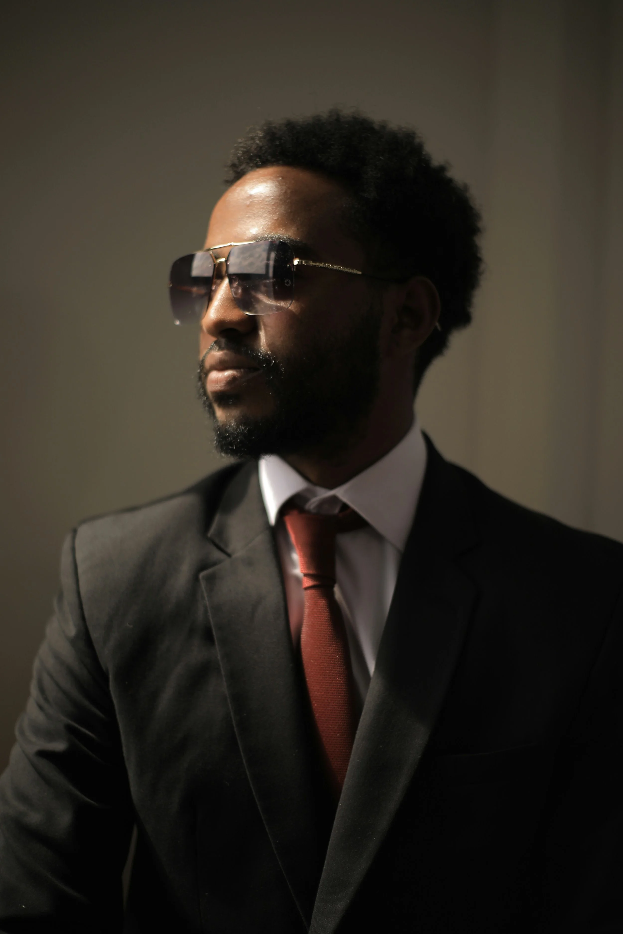 Man in black suit, white shirt, red tie, wearing sunglasses, with a short afro hairstyle and beard, against a plain background.