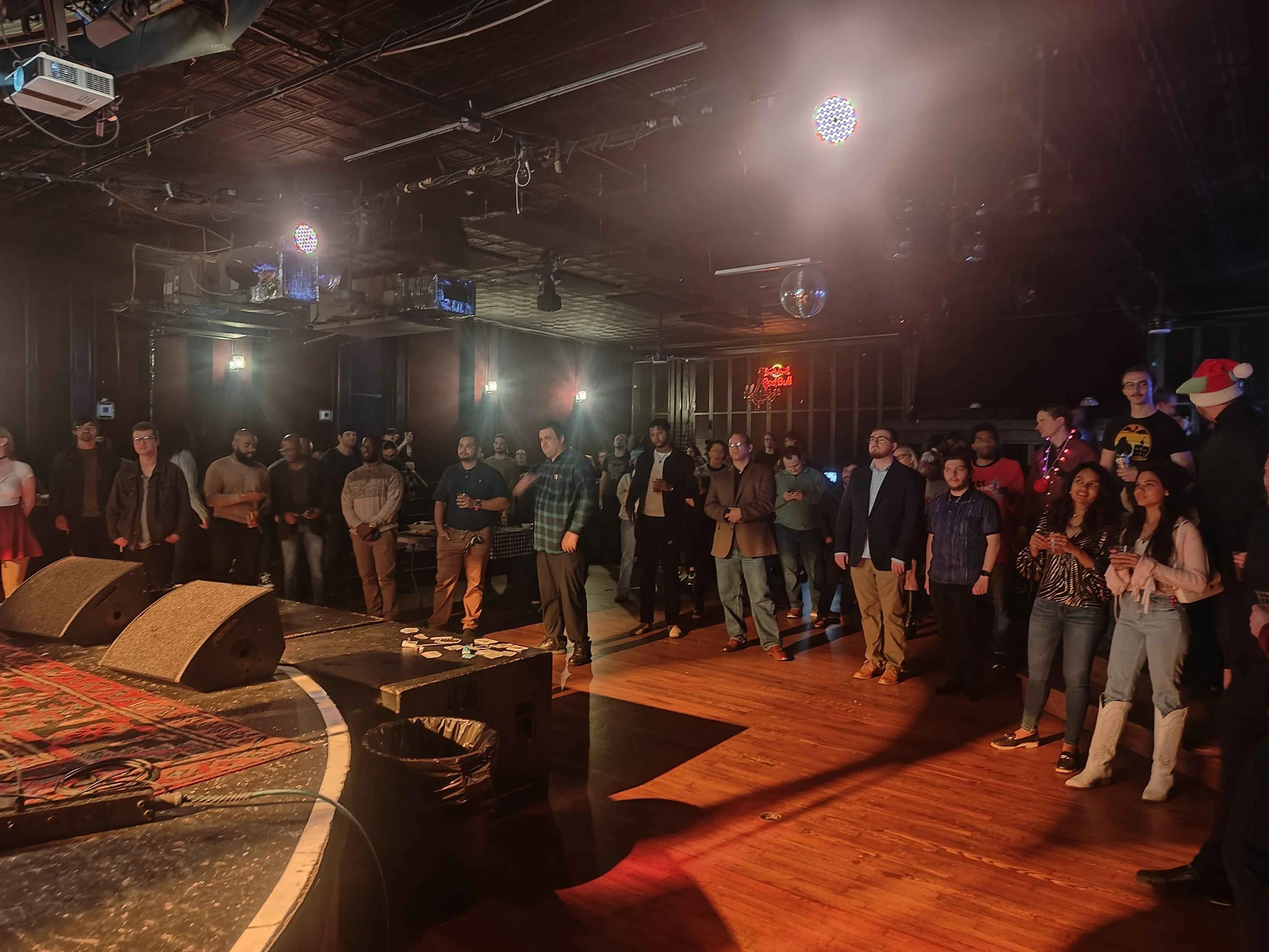 Crowd of people gathered in a dimly lit venue with wooden floors, stage speakers, and colorful lights, possibly at a concert or comedy event.