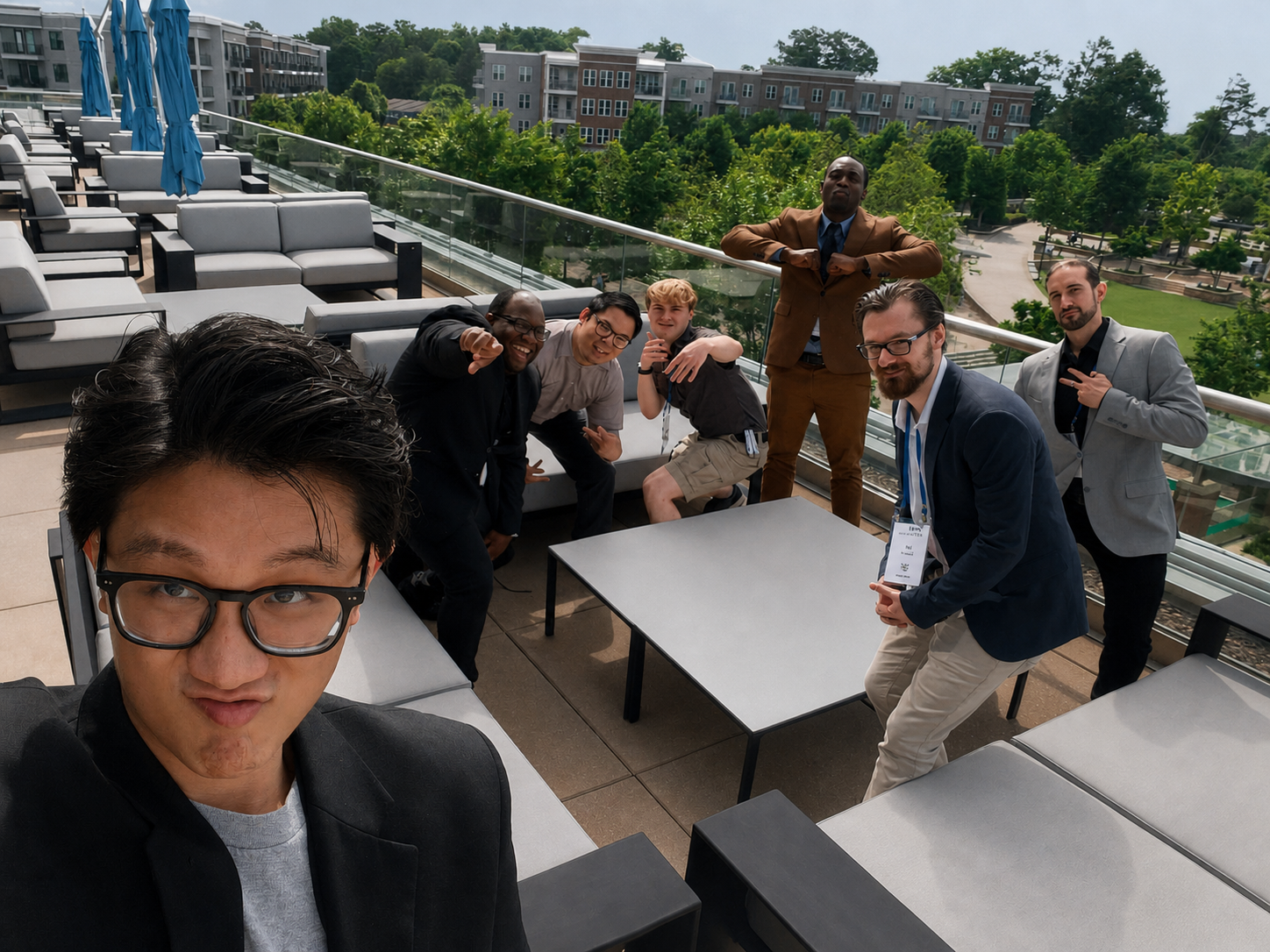 A group of seven men on a rooftop patio taking a selfie. They are smiling and posing with various gestures. The man in front with glasses is closest to the camera. The others are standing or crouching around a table, with a glass railing and trees with modern buildings in the background.
