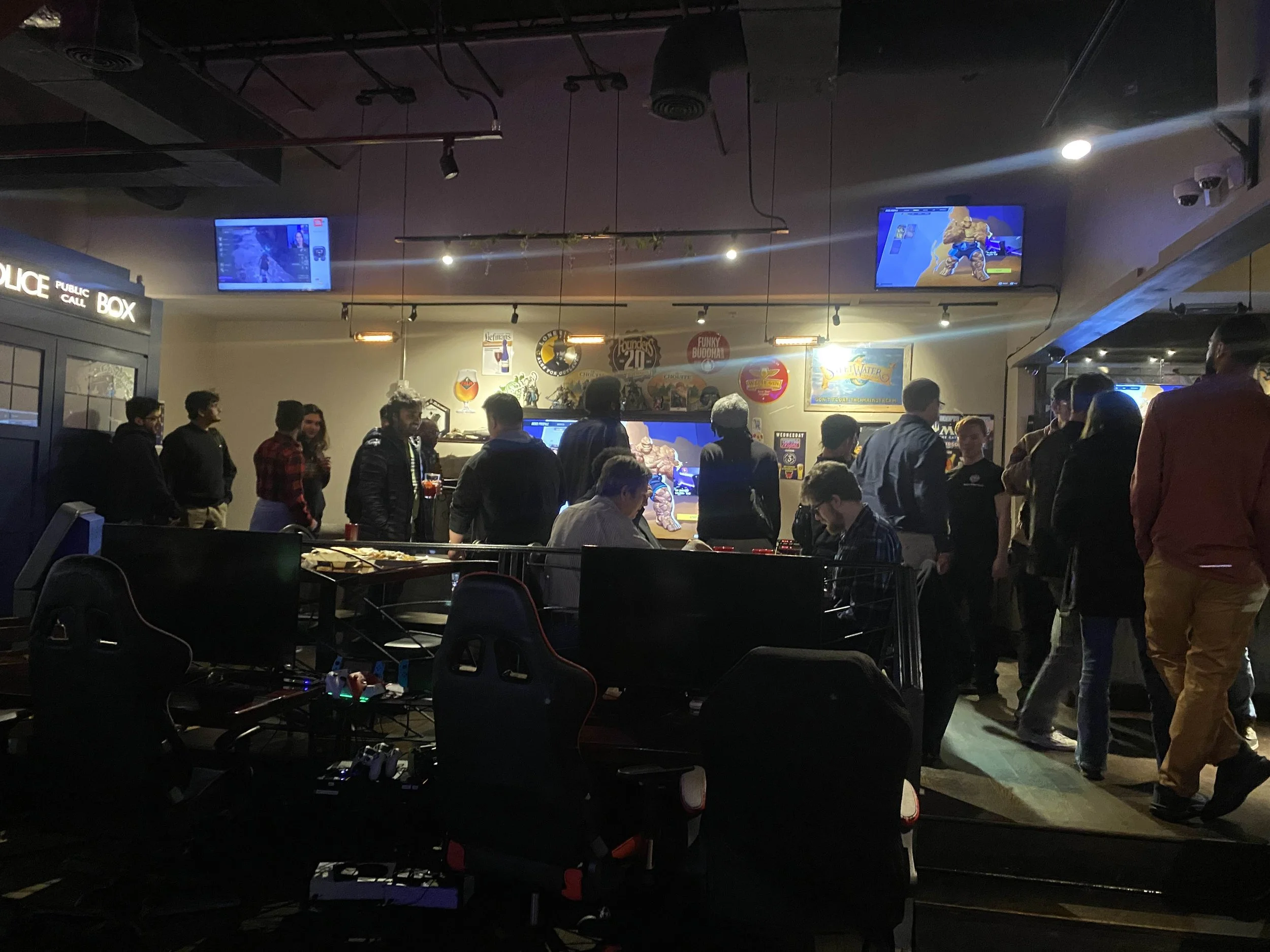 People gathered in a dimly lit bar or gaming venue watching large screens showing video games, with some individuals standing and some seated at gaming stations.