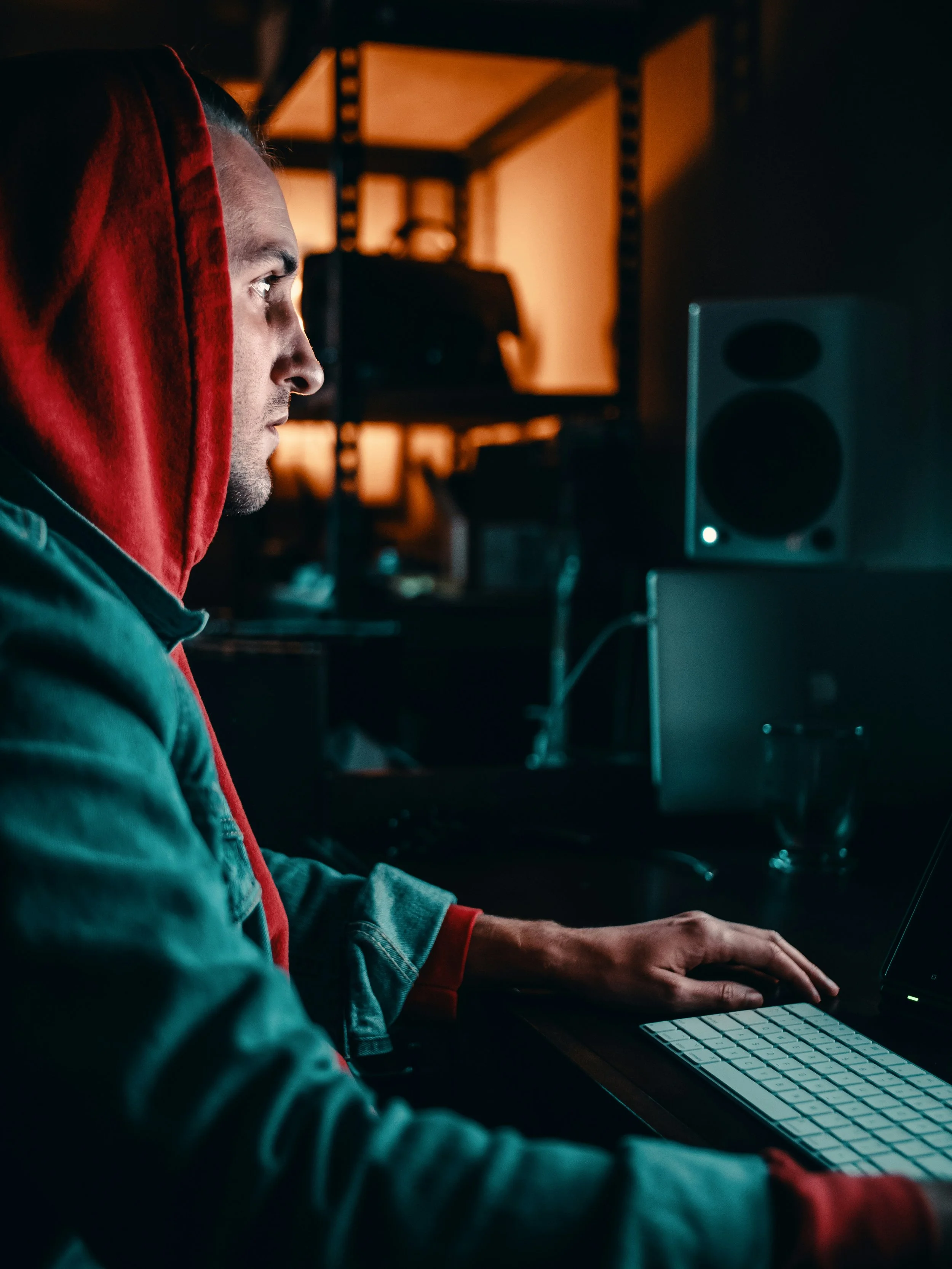 A person wearing a red hoodie and a denim jacket working on a computer in a dimly lit room with shelves in the background.