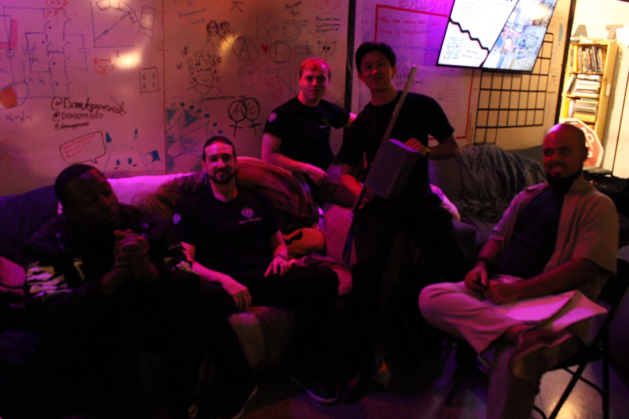 A group of five diverse young men sitting and standing in a dimly lit room with pink and purple lighting. The room has decorated whiteboards with drawings and writings, and a large TV screen mounted on the wall.