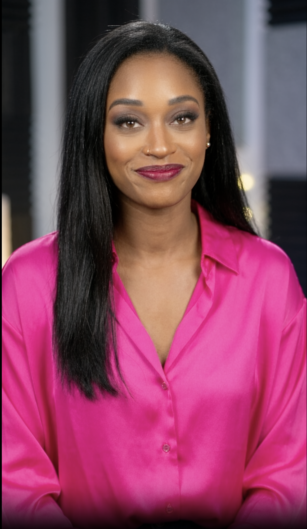 A woman with long black hair and medium skin tone wearing a bright pink satin shirt, smiling with dark makeup and lipstick, in an indoor setting.