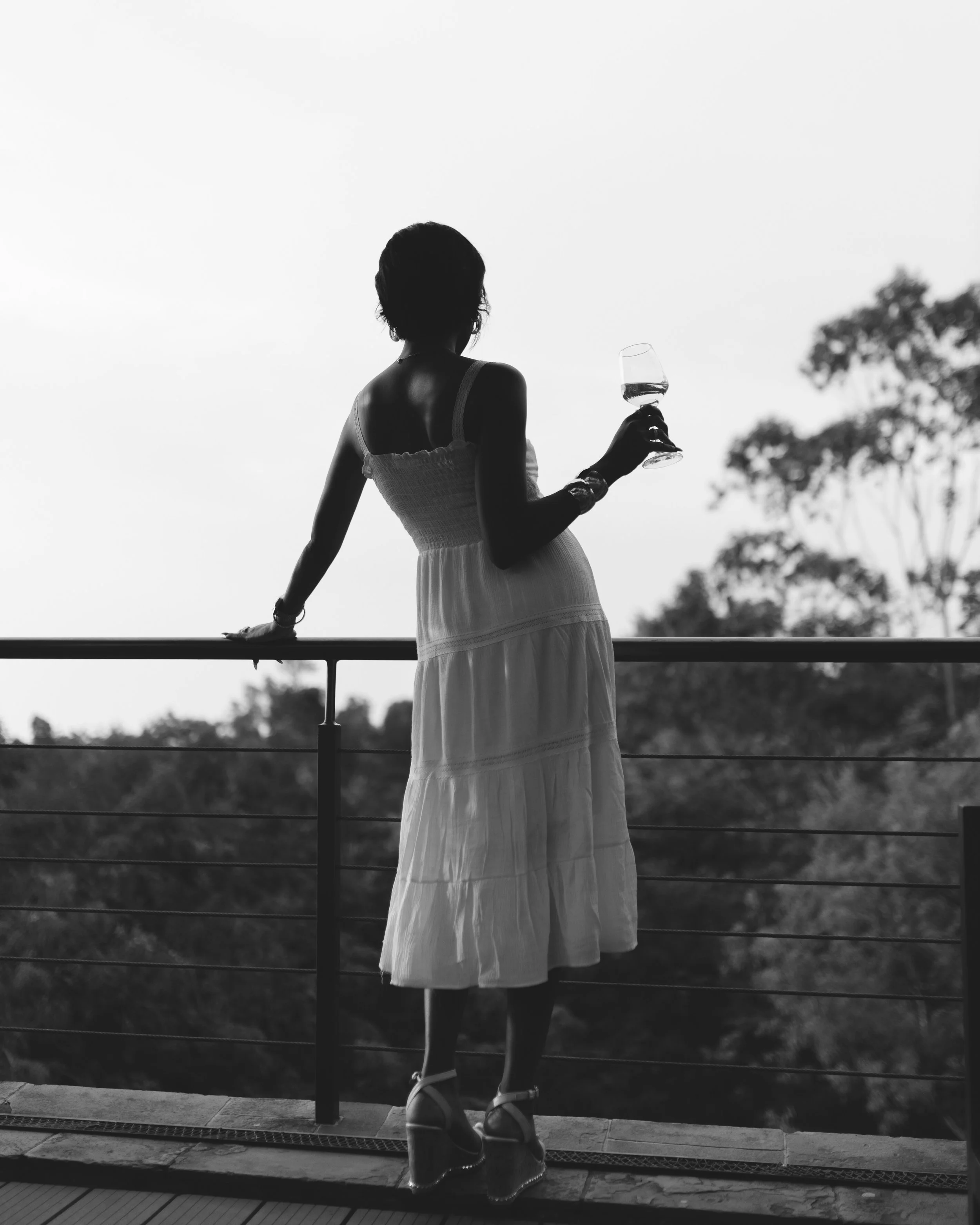 Black and white photo of a woman standing outdoors, holding a wine glass in her right hand, wearing a sleeveless dress and high-heeled sandals, with trees and sky in the background.