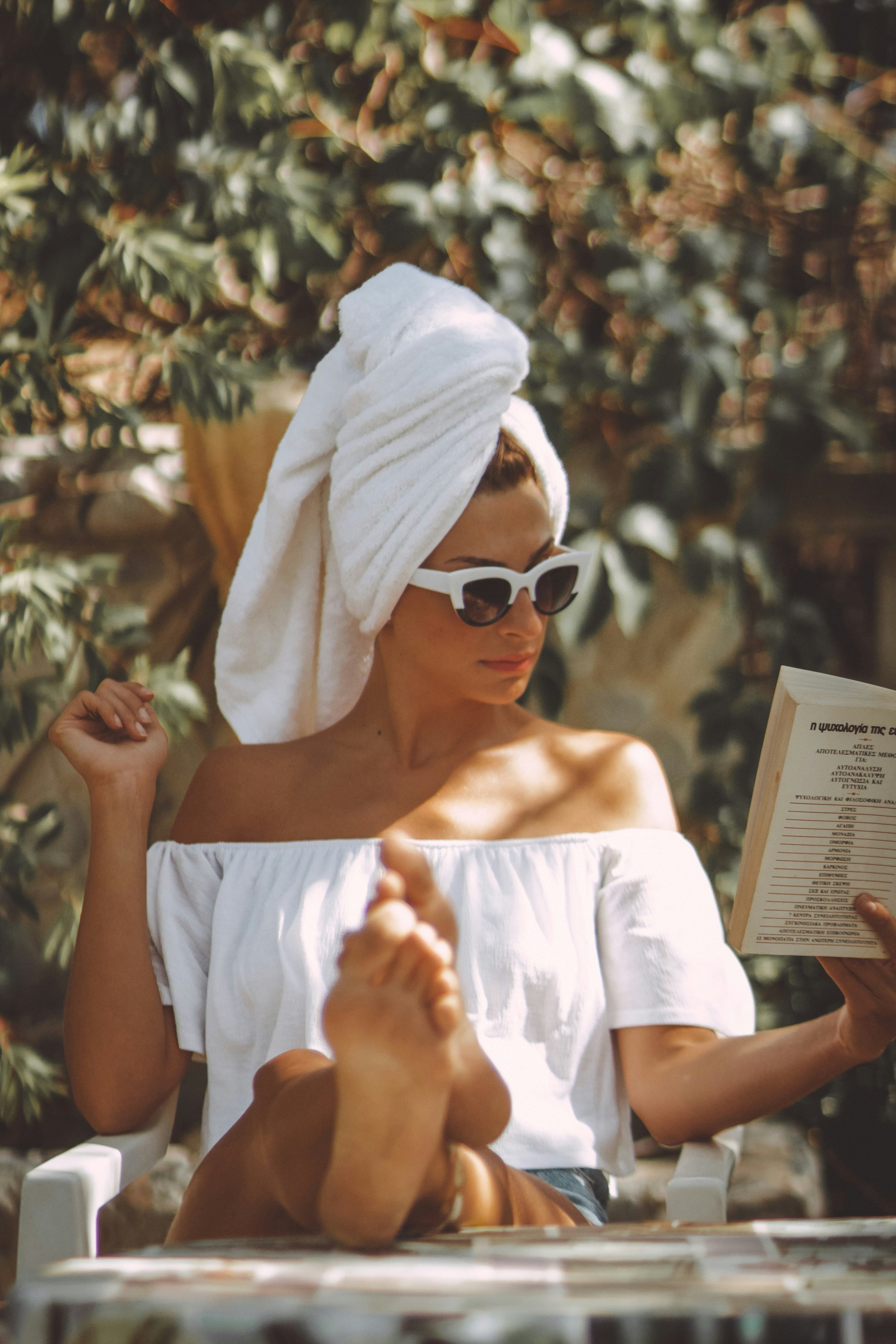 A woman with a towel wrapped around her head, wearing white sunglasses and an off-shoulder white top, reading a menu in an outdoor setting with a leafy background.
