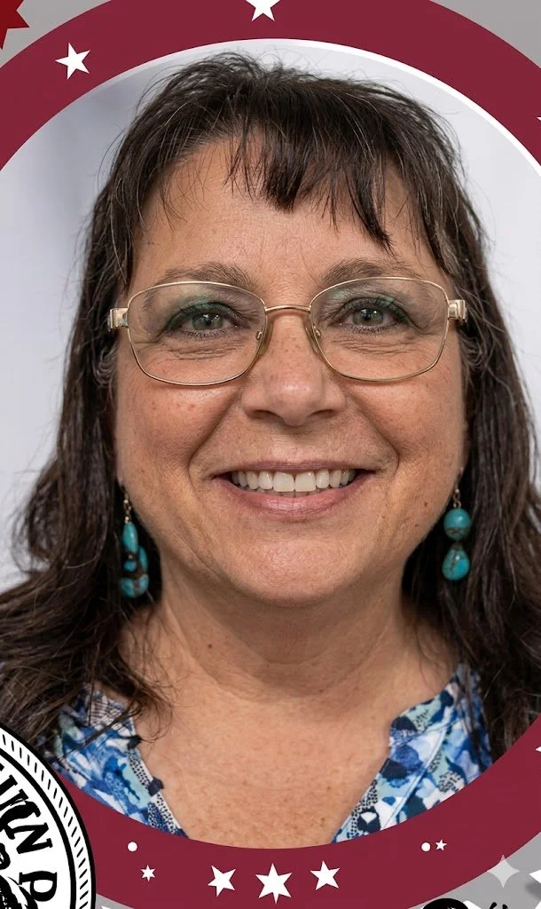A woman with glasses and dark brown hair smiling at the camera. She is wearing turquoise earrings and a blue patterned shirt, framed by a patriotic-themed border with stars.