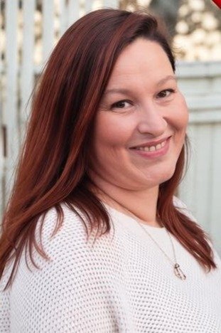 A woman with long red hair smiling outdoors, wearing a white sweater and a necklace, with a fence and trees in the background.