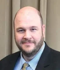 Portrait of a man with a bald head and beard, wearing a suit and tie, standing in front of beige curtains.