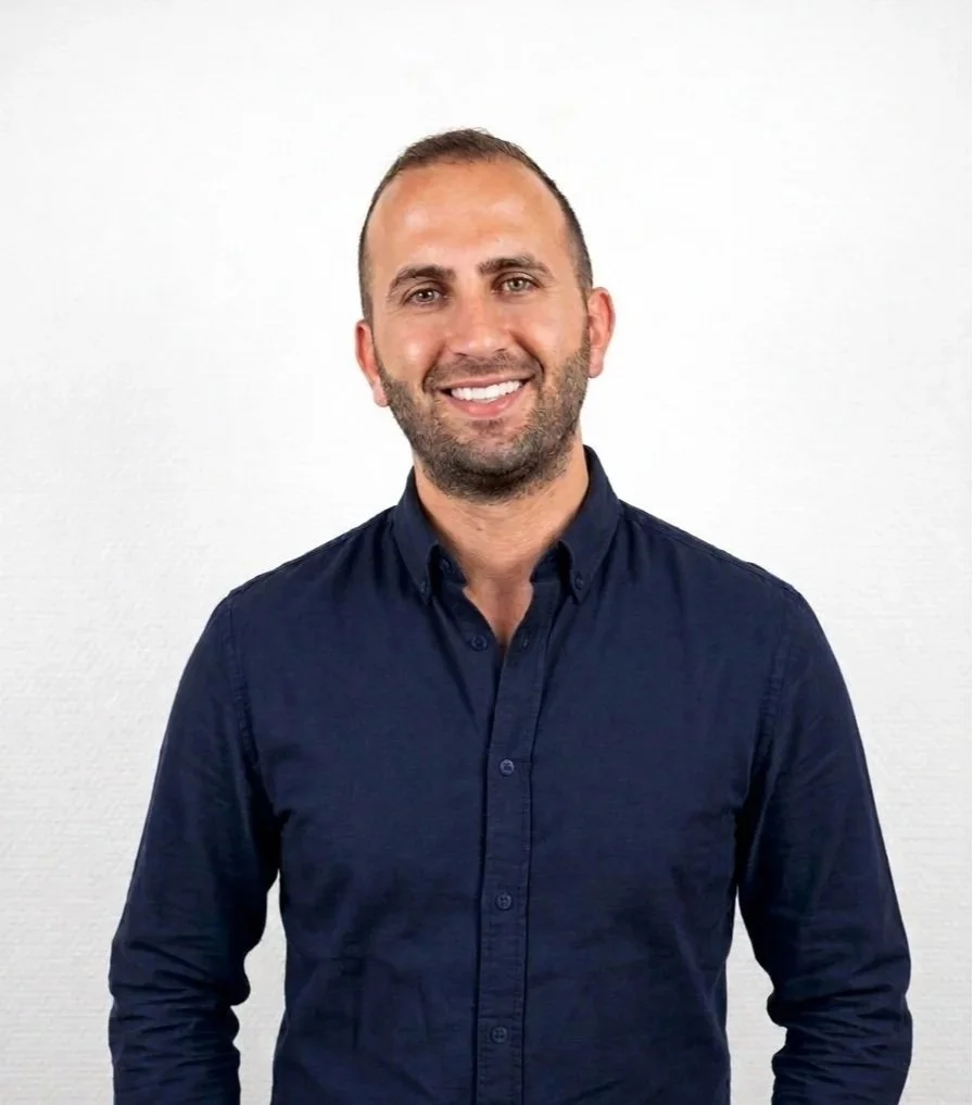 Smiling man with short hair and beard wearing a navy blue button-up shirt standing against a plain white background.