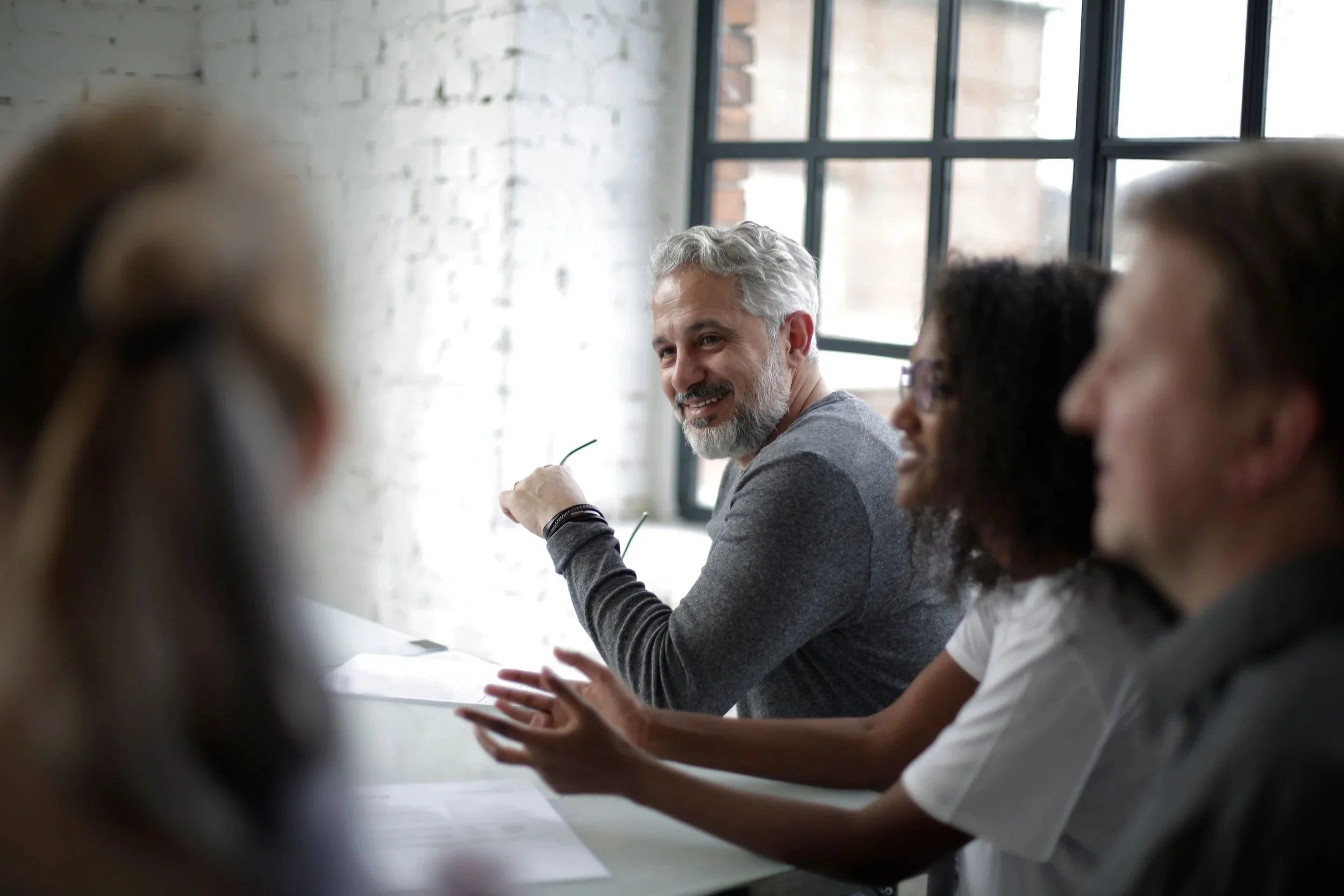 Group of people sitting at a table during a meeting in a modern office space with large windows and exposed brick wall, focusing on a middle-aged man with gray hair and a beard who is smiling and holding a pair of glasses.