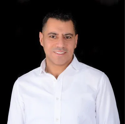 A man with short dark hair, wearing a white button-up shirt, smiling against a black background.