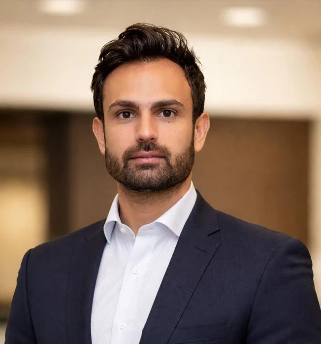 Portrait of a man with dark hair, beard, wearing a white shirt and dark blazer, standing indoors with blurred background.