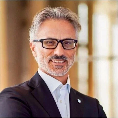 Portrait of a middle-aged man with gray hair and beard, wearing glasses and a suit, standing indoors with a blurred background.