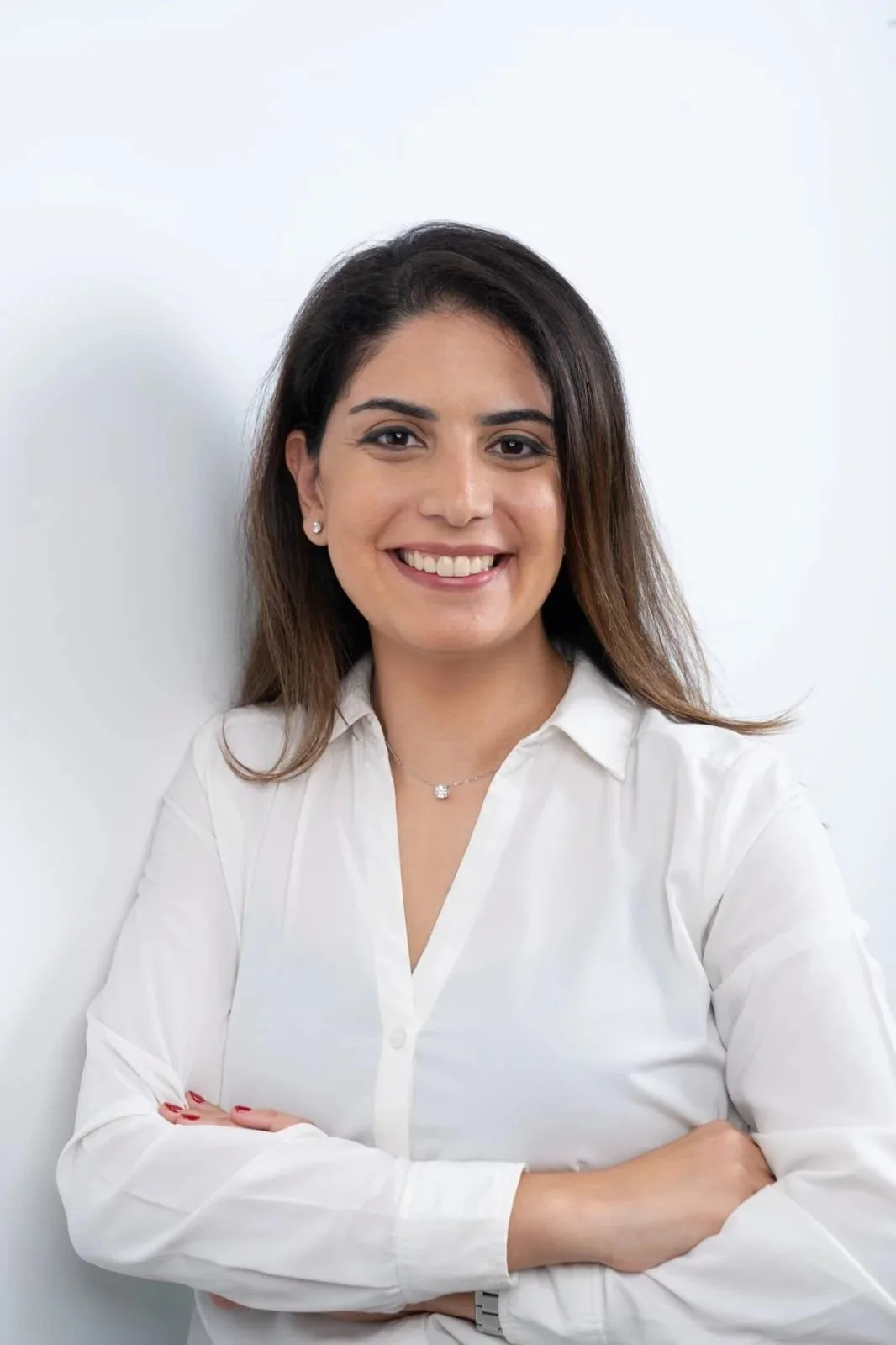 Portrait of a young woman with brown hair, smiling, wearing a white shirt and jewelry, standing against a white background.