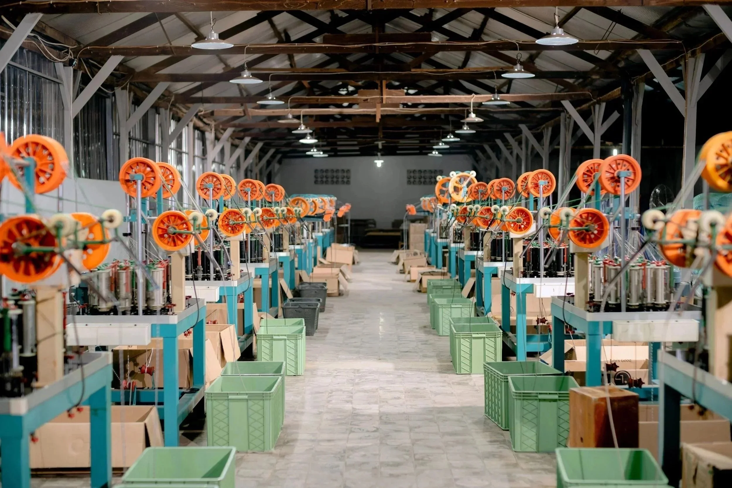 A large indoor space with rows of machinery with orange wheels and white and blue structural frames, and green plastic bins on the floor, under a high ceiling with exposed wooden beams and hanging lights.