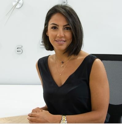 Woman with shoulder-length dark hair sitting at a desk in an office, smiling, against a white wall with a clock.