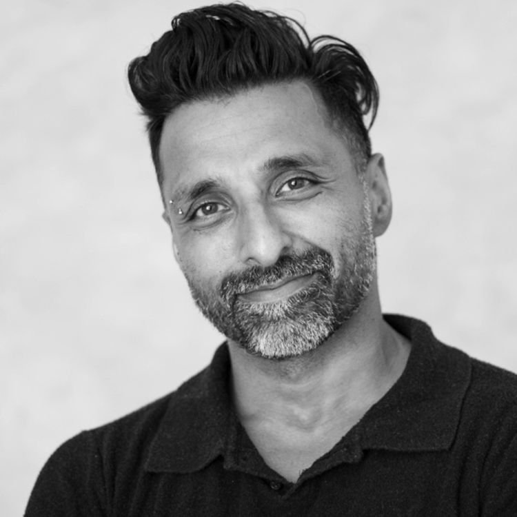 Black and white portrait of a man with short, styled hair, a beard, and a small piercing above his right eyebrow, smiling slightly, wearing a dark collared shirt.