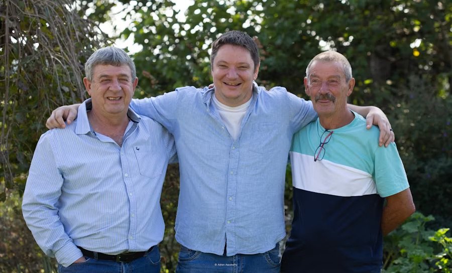 Three men standing outside with arms around each other, smiling in front of green trees.