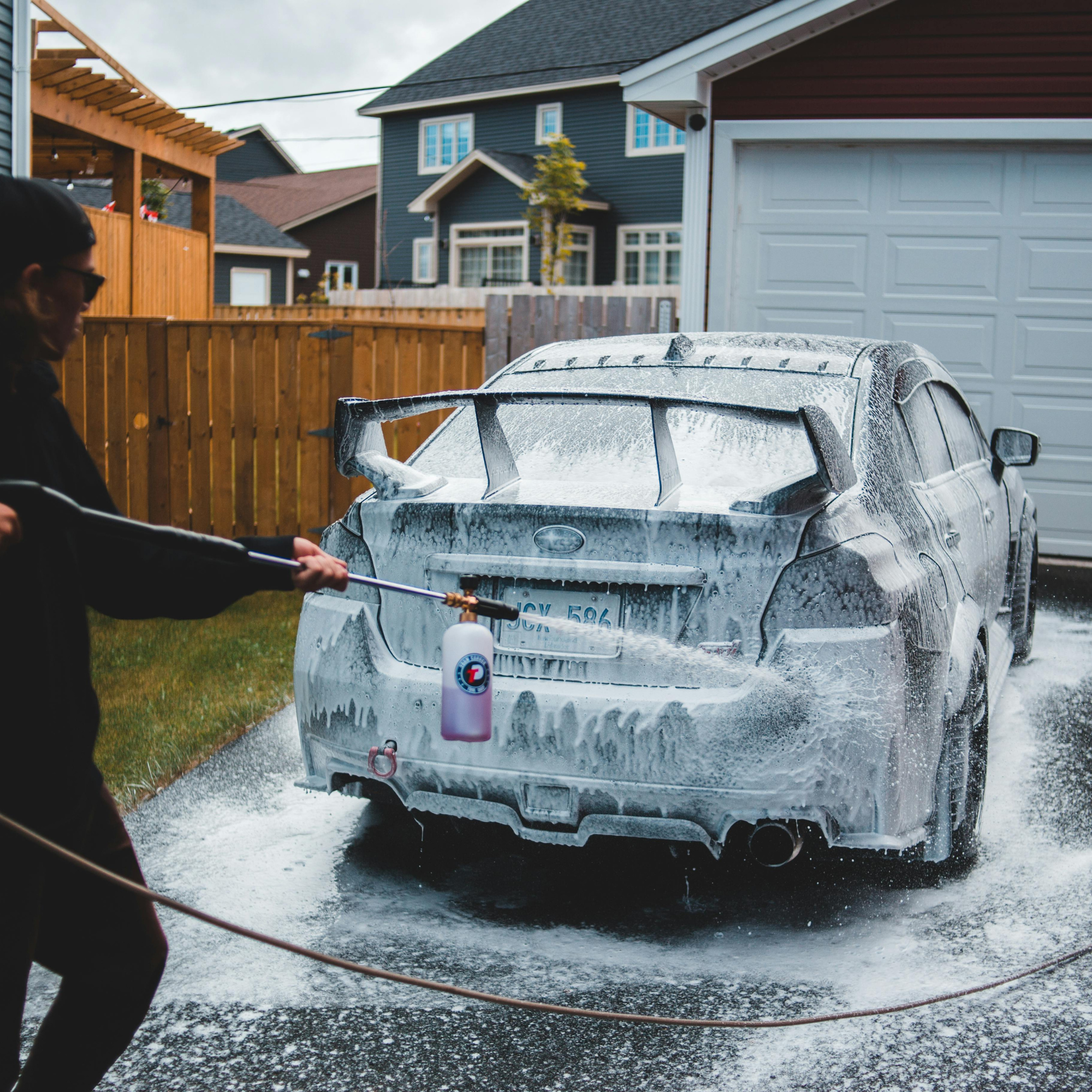 A person holding a pressure washer with soap foam, washing a car in a driveway in front of a garage, with residential houses in the background.