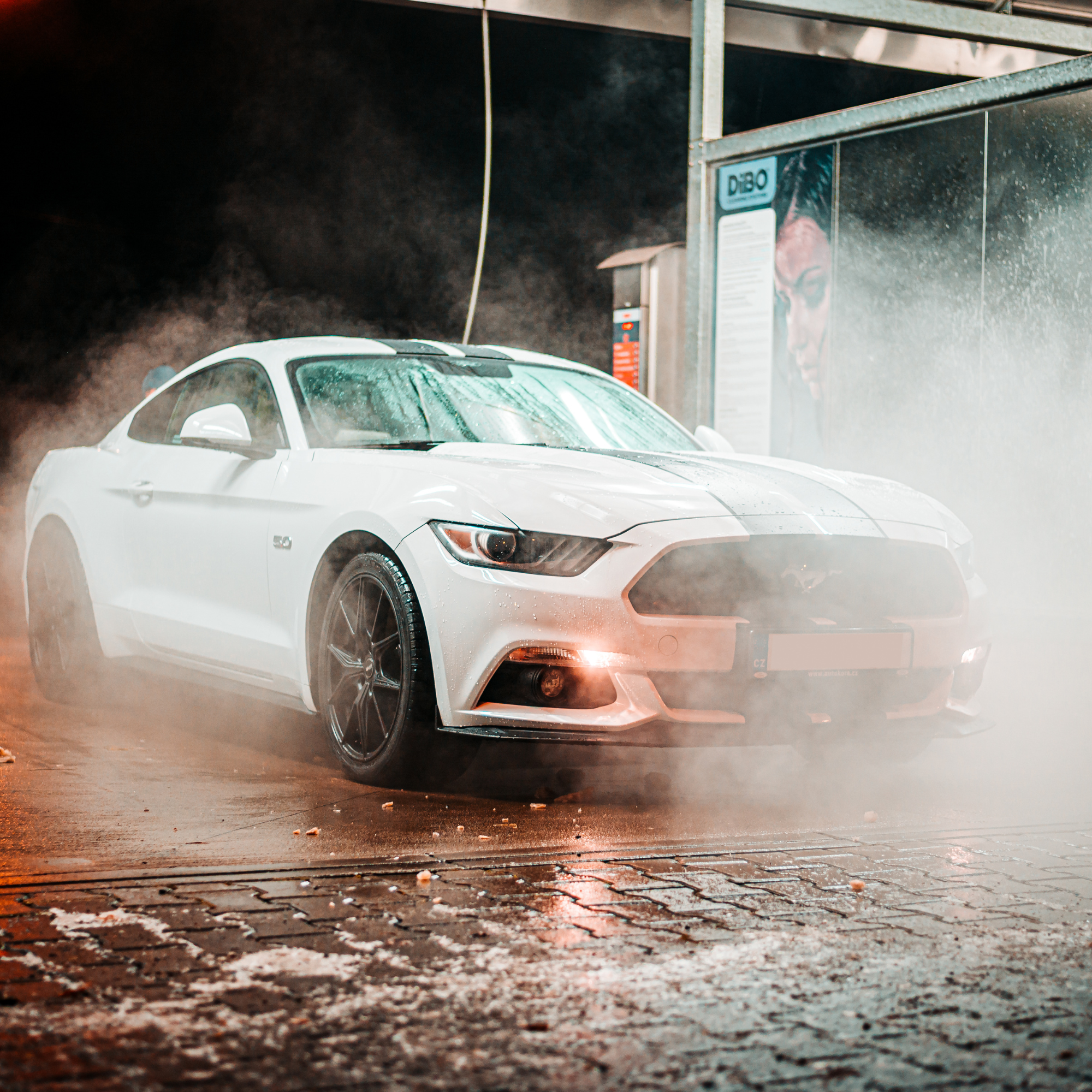 A white sports car washing at an automatic car wash with water spray and soap foam, on a wet asphalt ground with scattered debris.