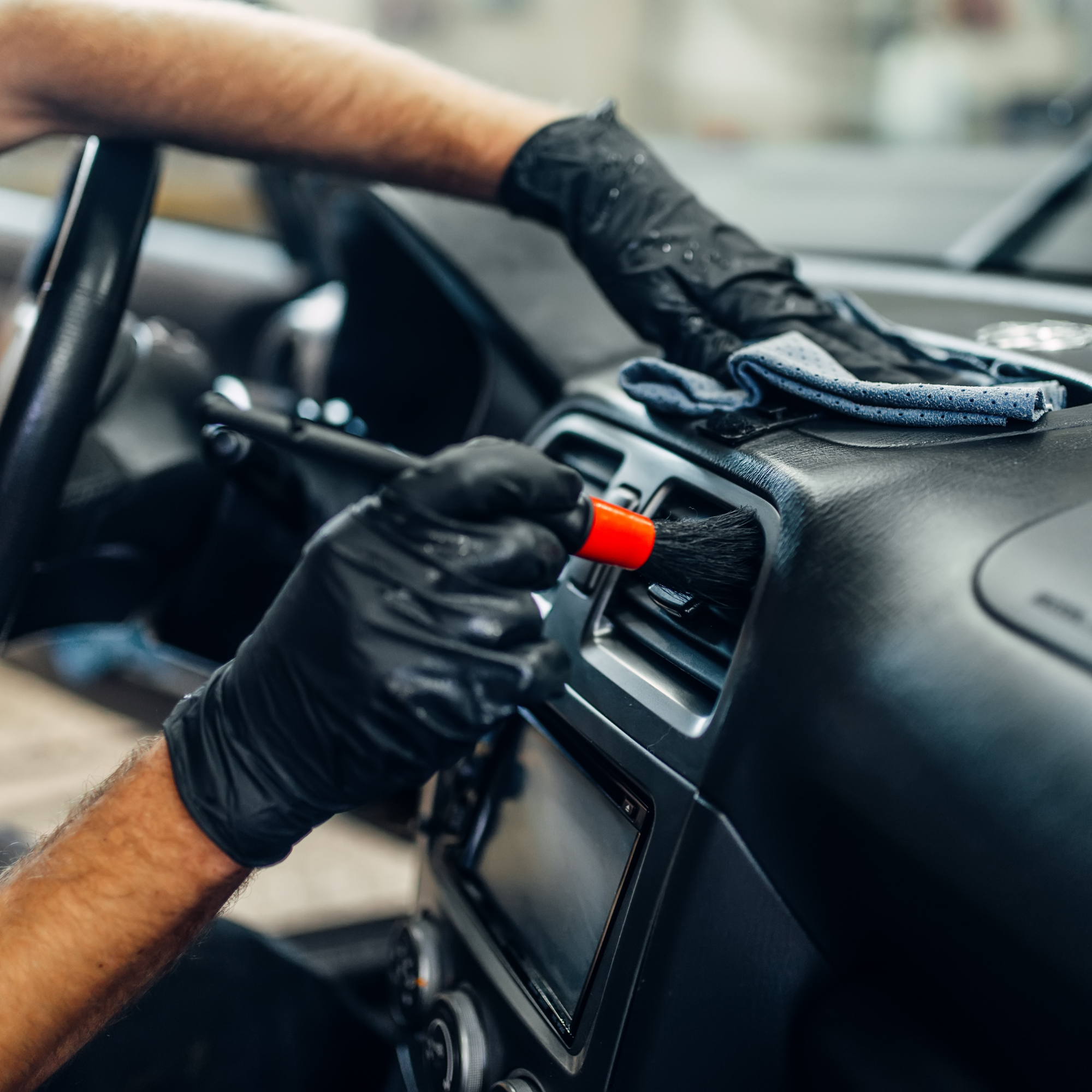A person's arm wearing a black glove is cleaning or adjusting the dashboard of a car with a cloth. The image focuses on the arm and the area around the dashboard.