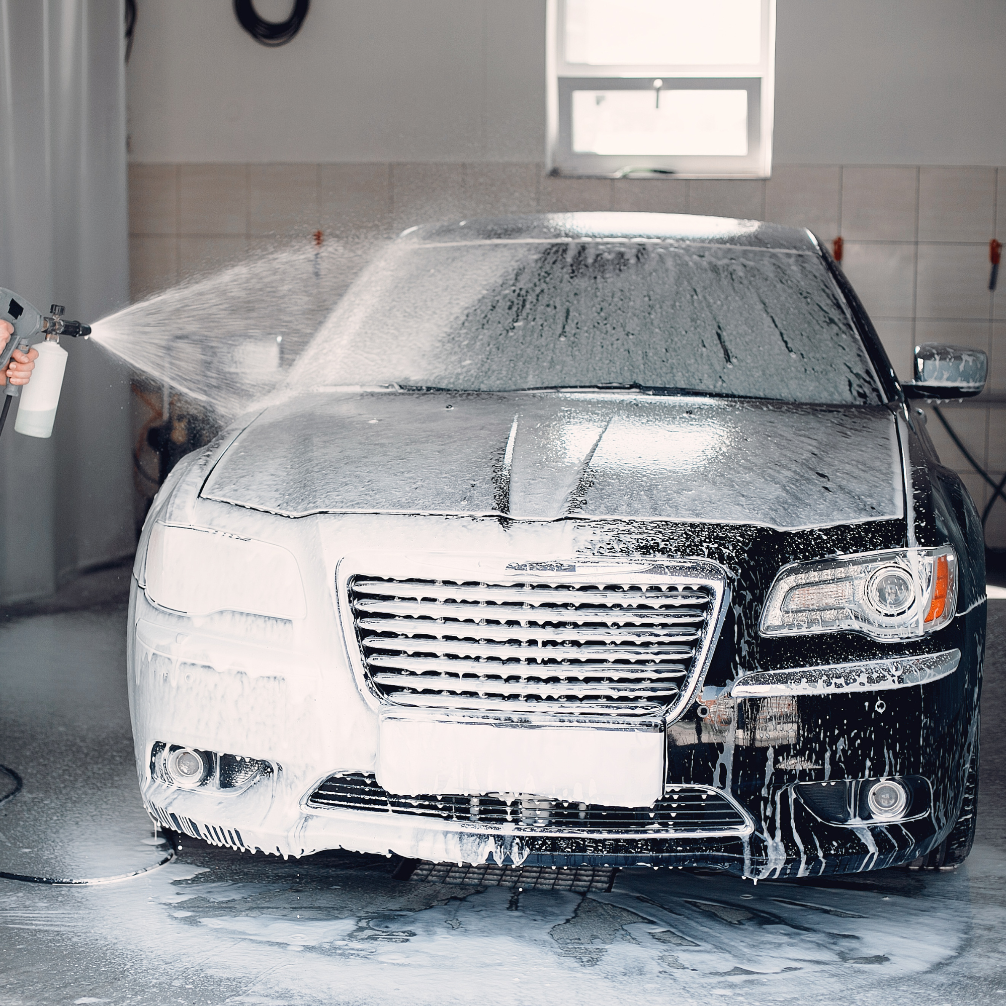 A black car is being washed with foam. The front of the car, including the grille and headlights, is visible. A person is spraying foam onto the car in a garage or car wash area.