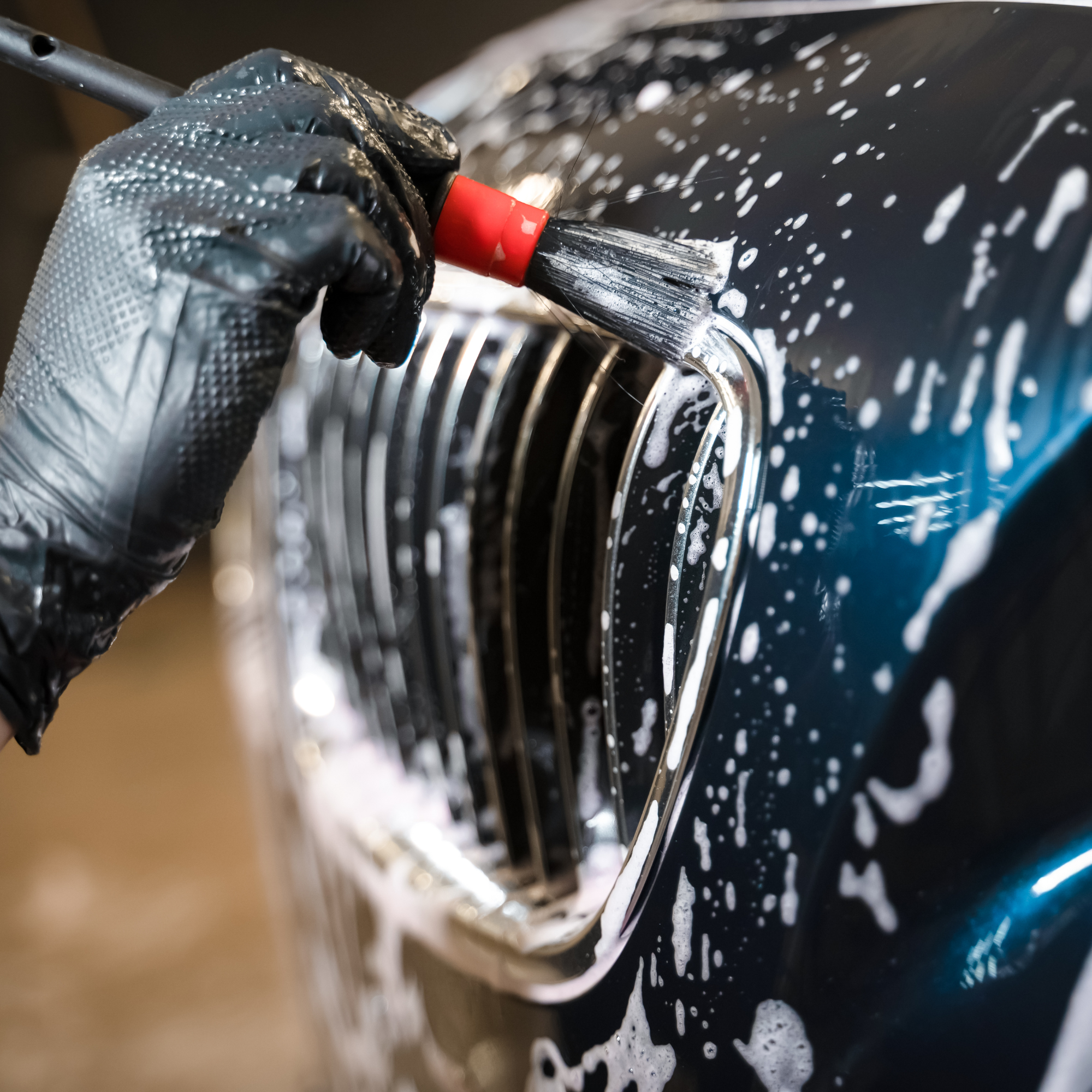 Close-up of a person's hand wearing a gray textured glove, cleaning or polishing a black surface with a small red and black brush with soap suds and white foam.