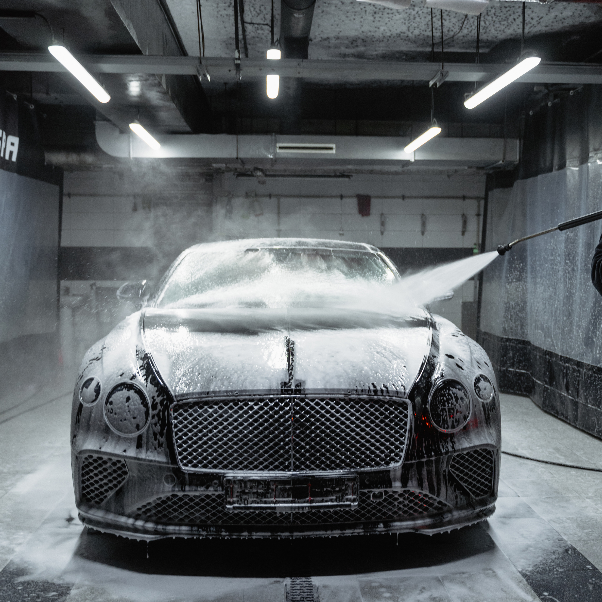 A luxury black car, likely a Mercedes-Benz, is being washed with soap and water in an indoor car wash station. The front of the car features distinctive headlights and grille, with water spray and soap suds visible.