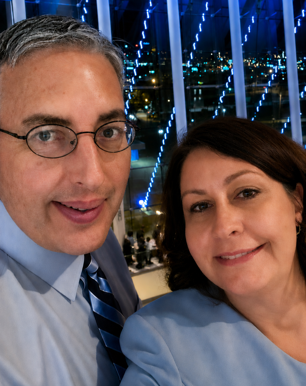 Two people taking a selfie at night with city lights and blue decorative lights in the background.