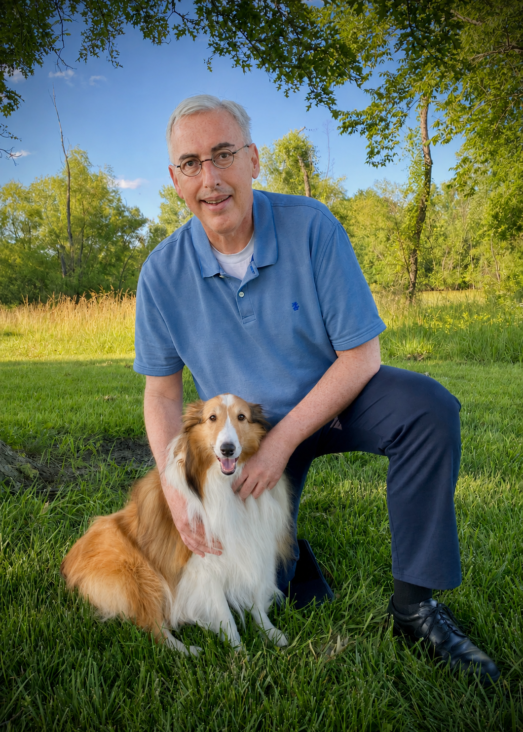 A man with glasses wearing a blue polo shirt crouches in a grassy park with a friendly, happy dog, surrounded by trees and a clear blue sky.