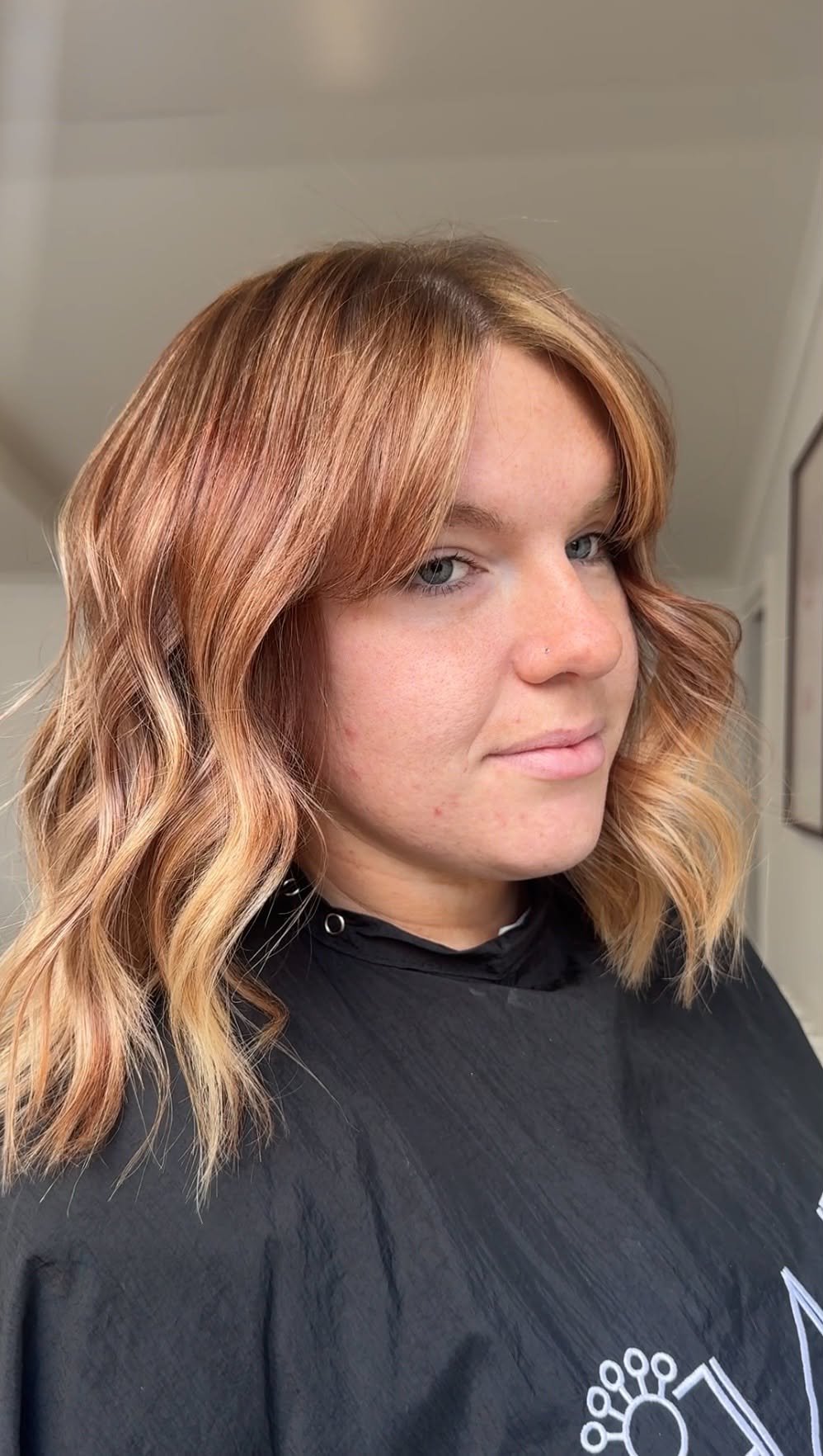 A woman with shoulder-length wavy hair dyed in shades of auburn and blonde, wearing a black cape, smiling slightly, in a salon setting.