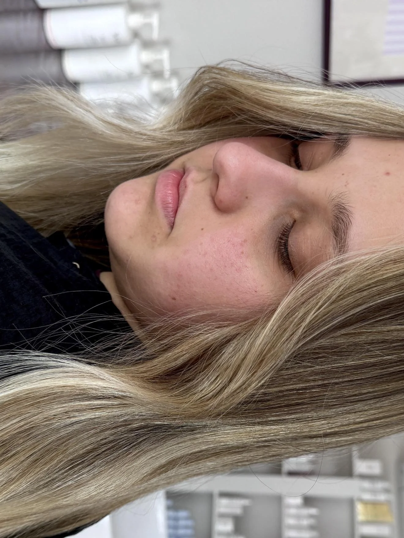 A young woman with long blonde hair and light skin is resting with her eyes closed. The background shows a blurred view of an indoor environment, possibly an office or classroom, with white shelves and papers.