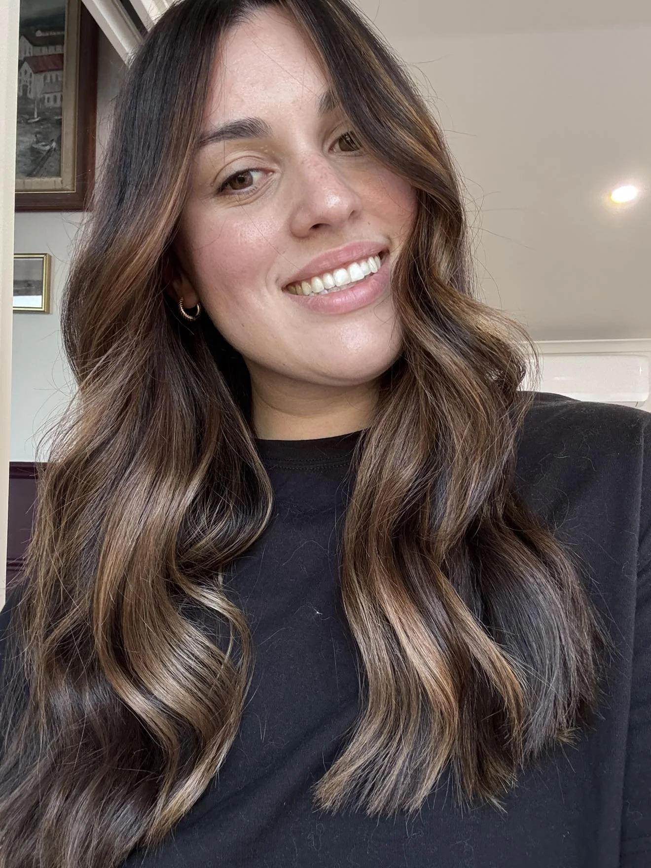 A smiling young woman with long, wavy brown hair and light skin, wearing a black top and small hoop earrings, taking a selfie indoors with picture frames on the wall in the background.