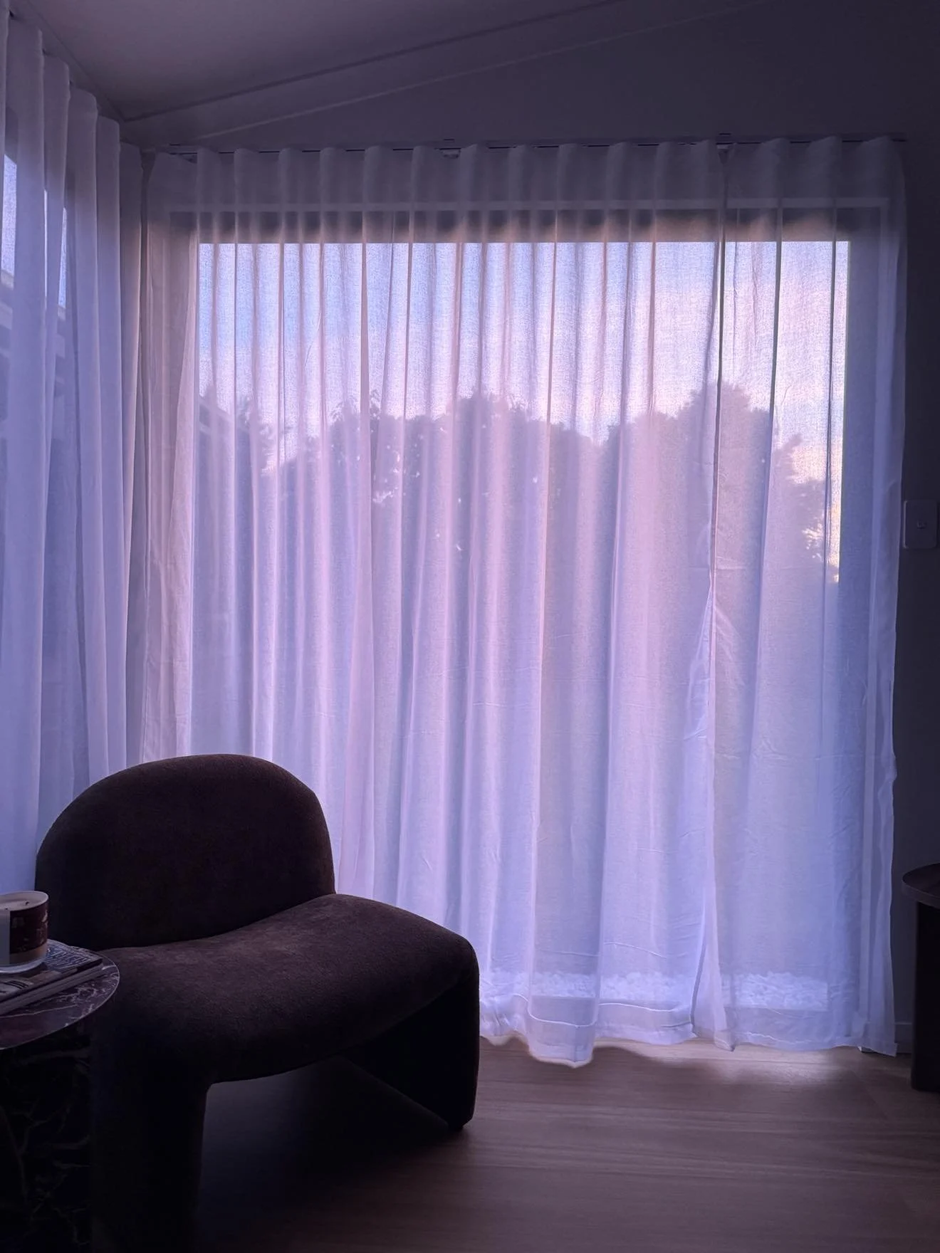 A room with a large window covered by white sheer curtains, with trees visible outside during sunset. There is a dark-colored armchair partially visible in the lower left corner and a small side table with objects on it.