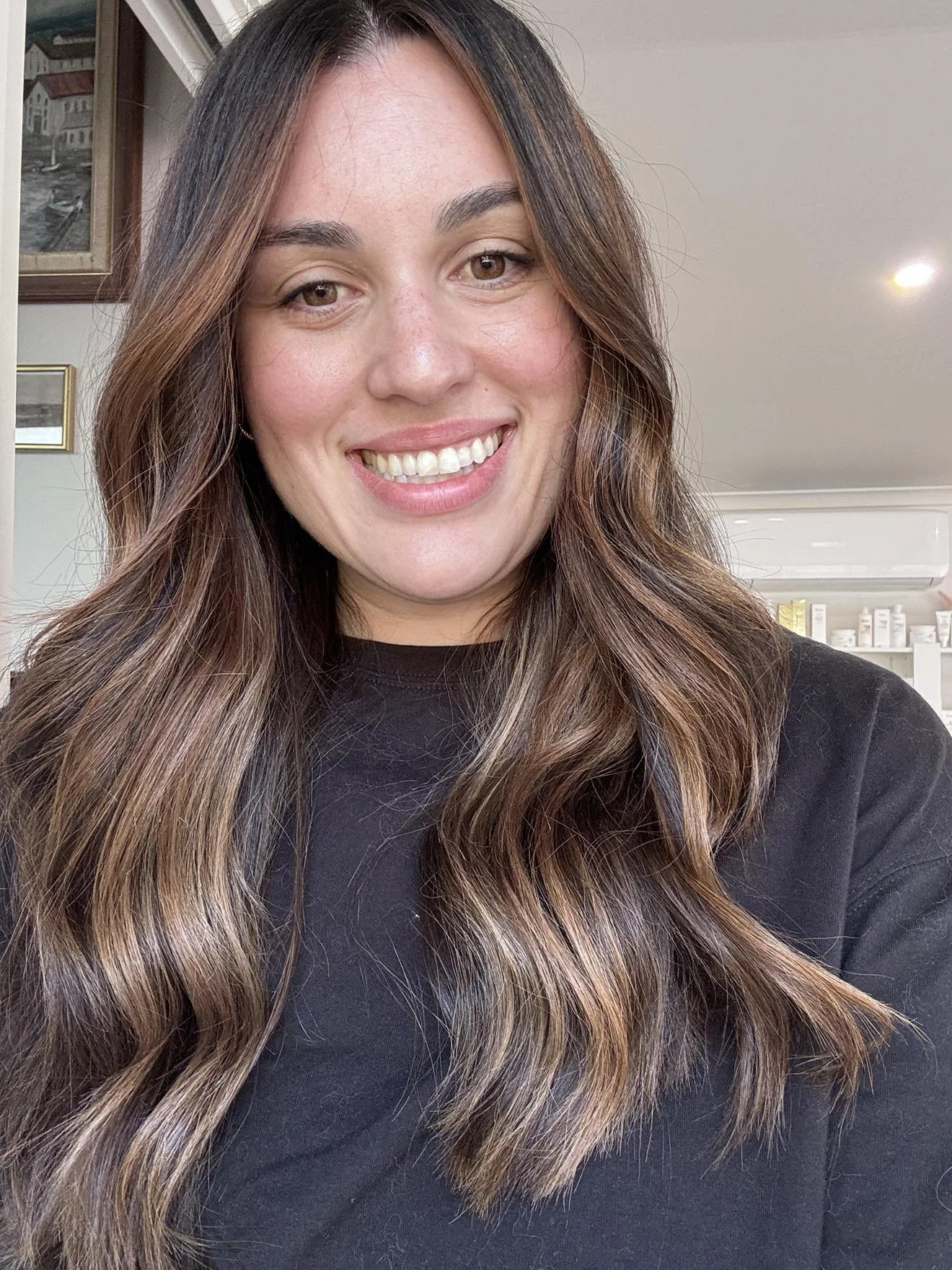 A woman with long, wavy brown hair with highlights, smiling, wearing a black top, in a room with framed pictures and a white air conditioning unit in the background.