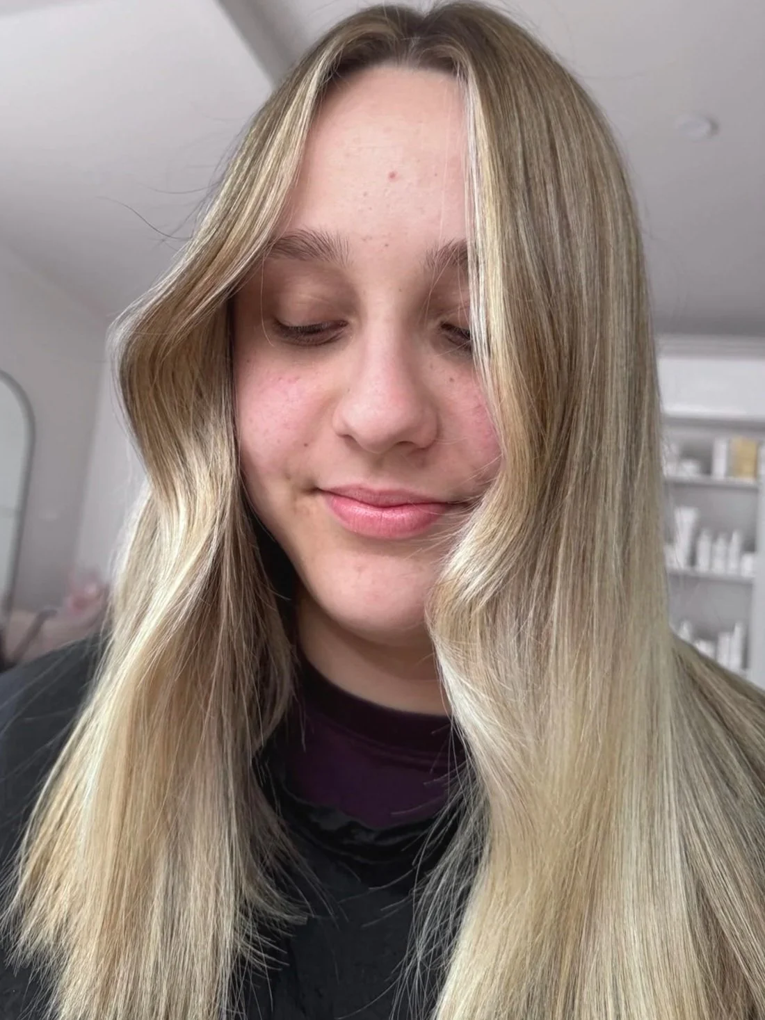 A young woman with long blonde hair, closed eyes, and a slight smile, in an indoor setting with shelves and products in the background.