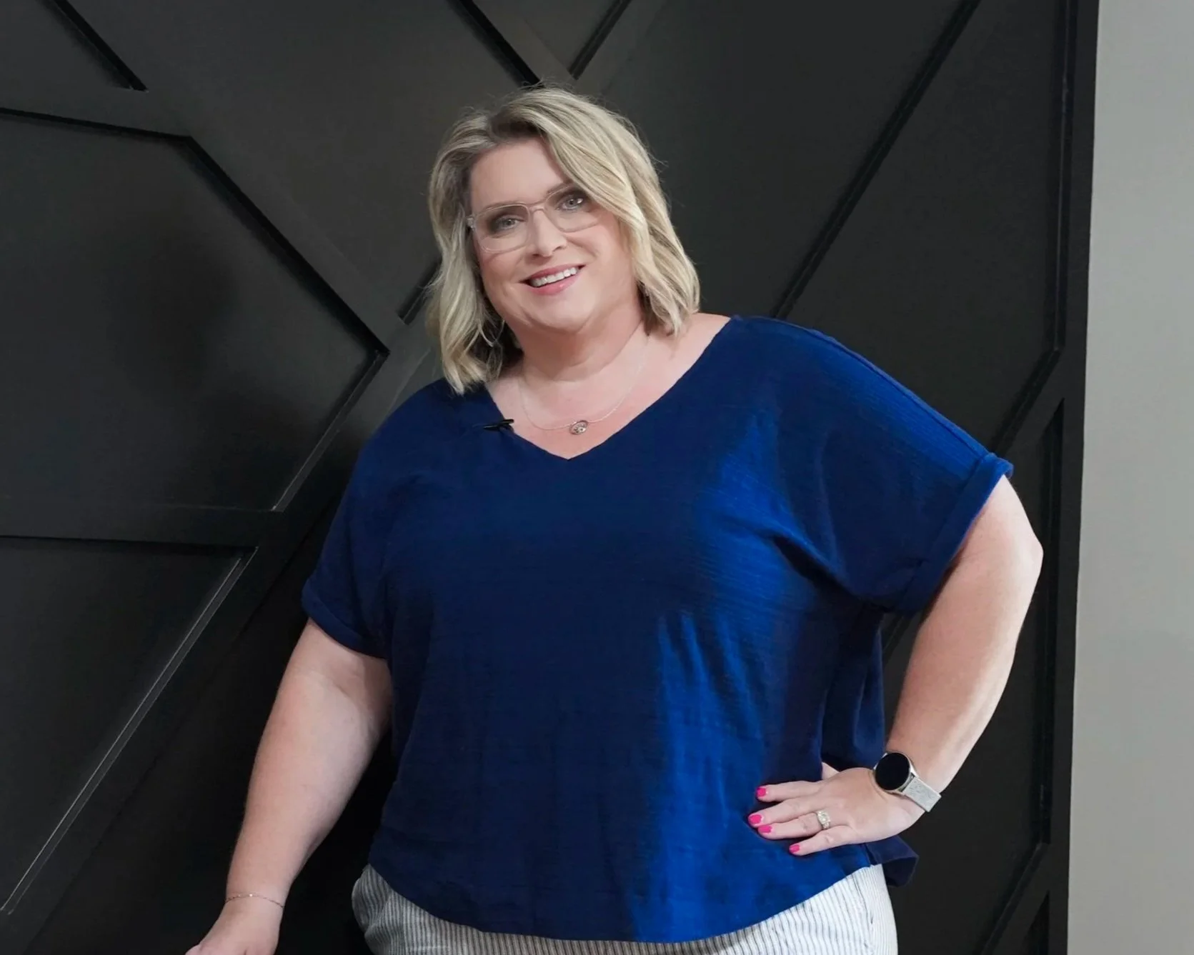 A woman wearing glasses, a blue top, and striped pants posing with her hand on her hip in front of a black geometric wall.
