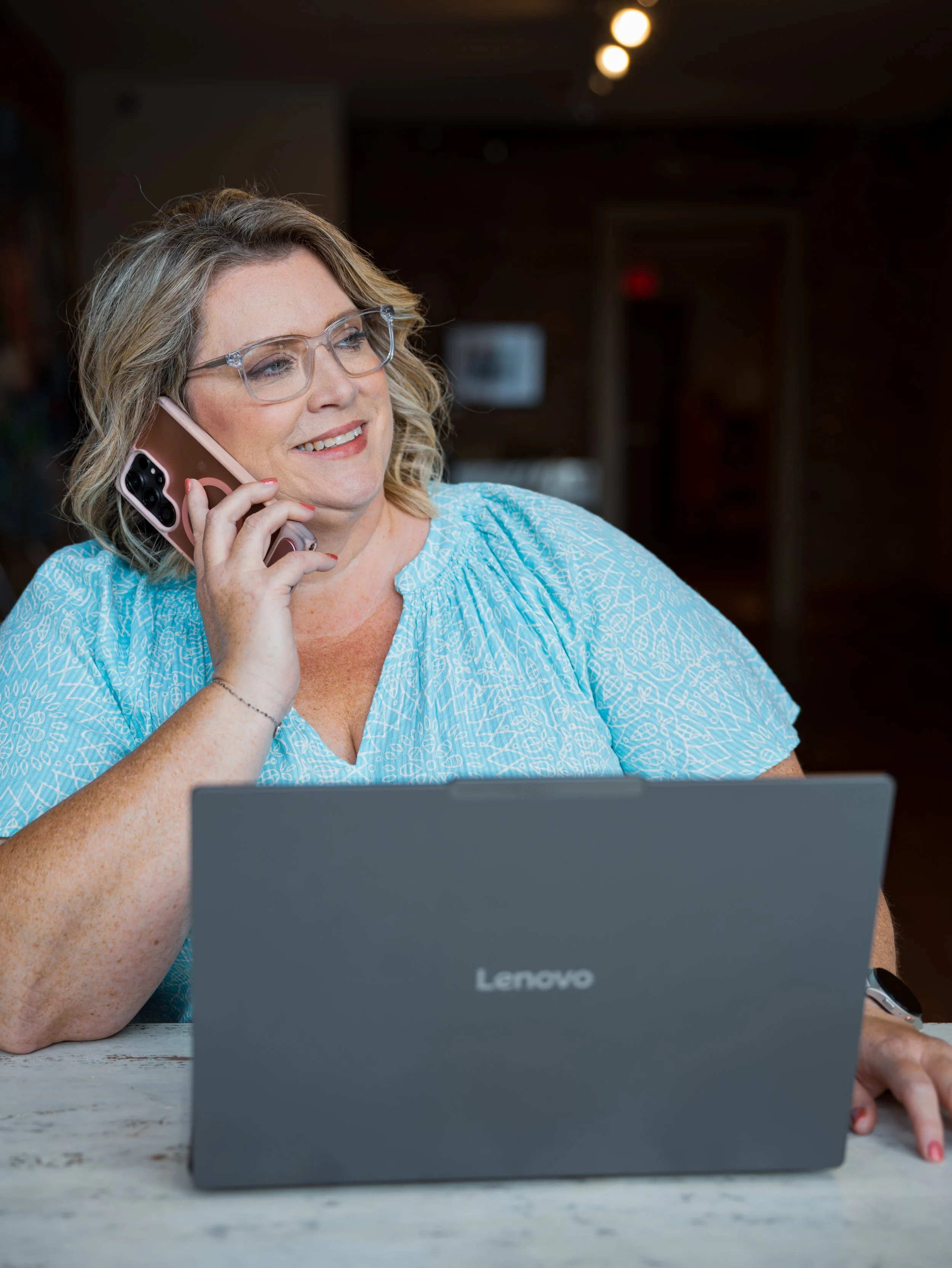 A smiling woman with blonde, curly hair wearing glasses, a blue patterned blouse, and a thin bracelet talking on a pink phone. She is sitting at a table in front of a gray Lenovo laptop. The background is dimly lit.