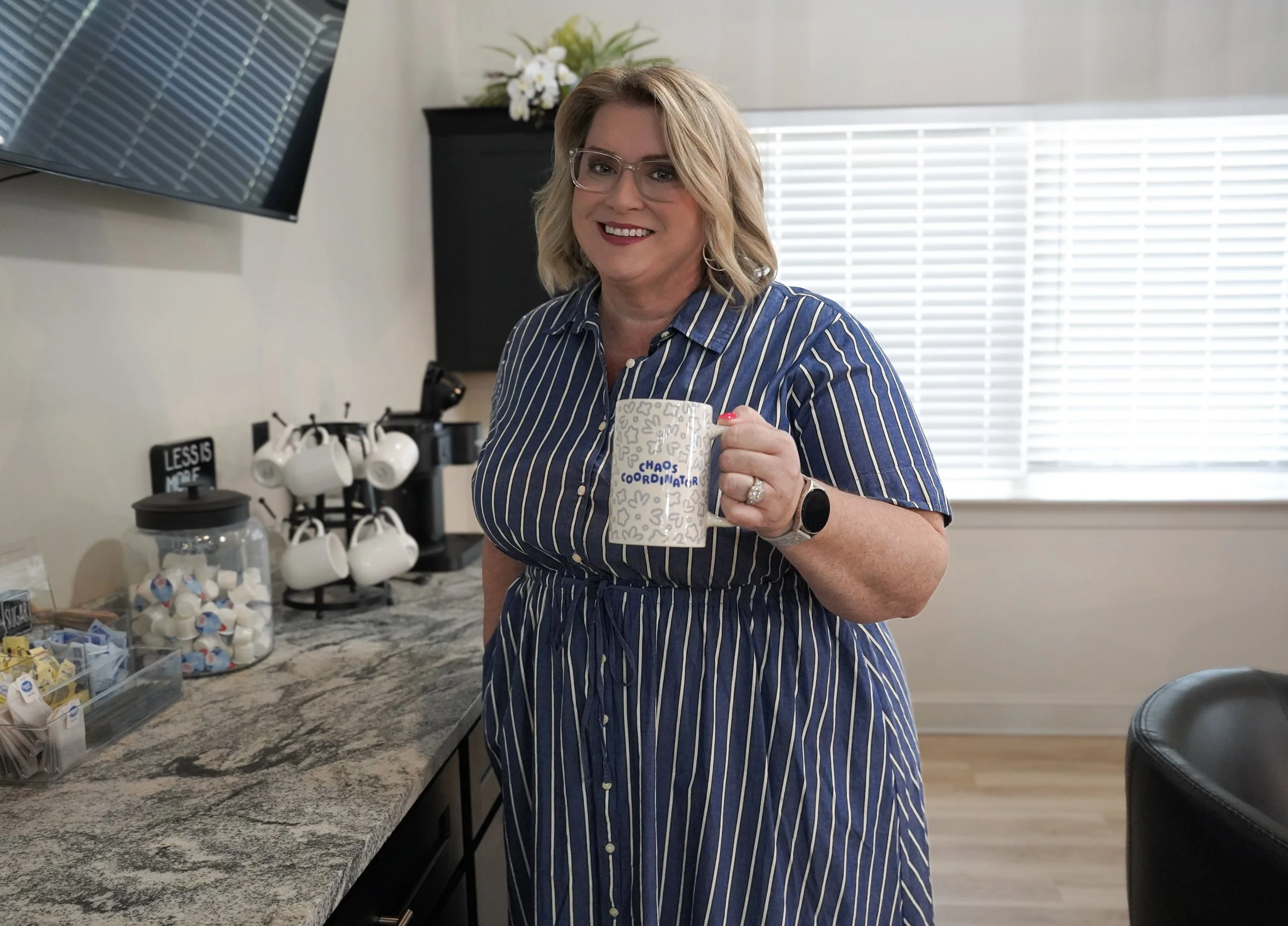 A woman with blonde hair, wearing glasses and a blue striped dress, smile while holding a coffee mug in a kitchen with coffee pods and a coffee maker on the counter.