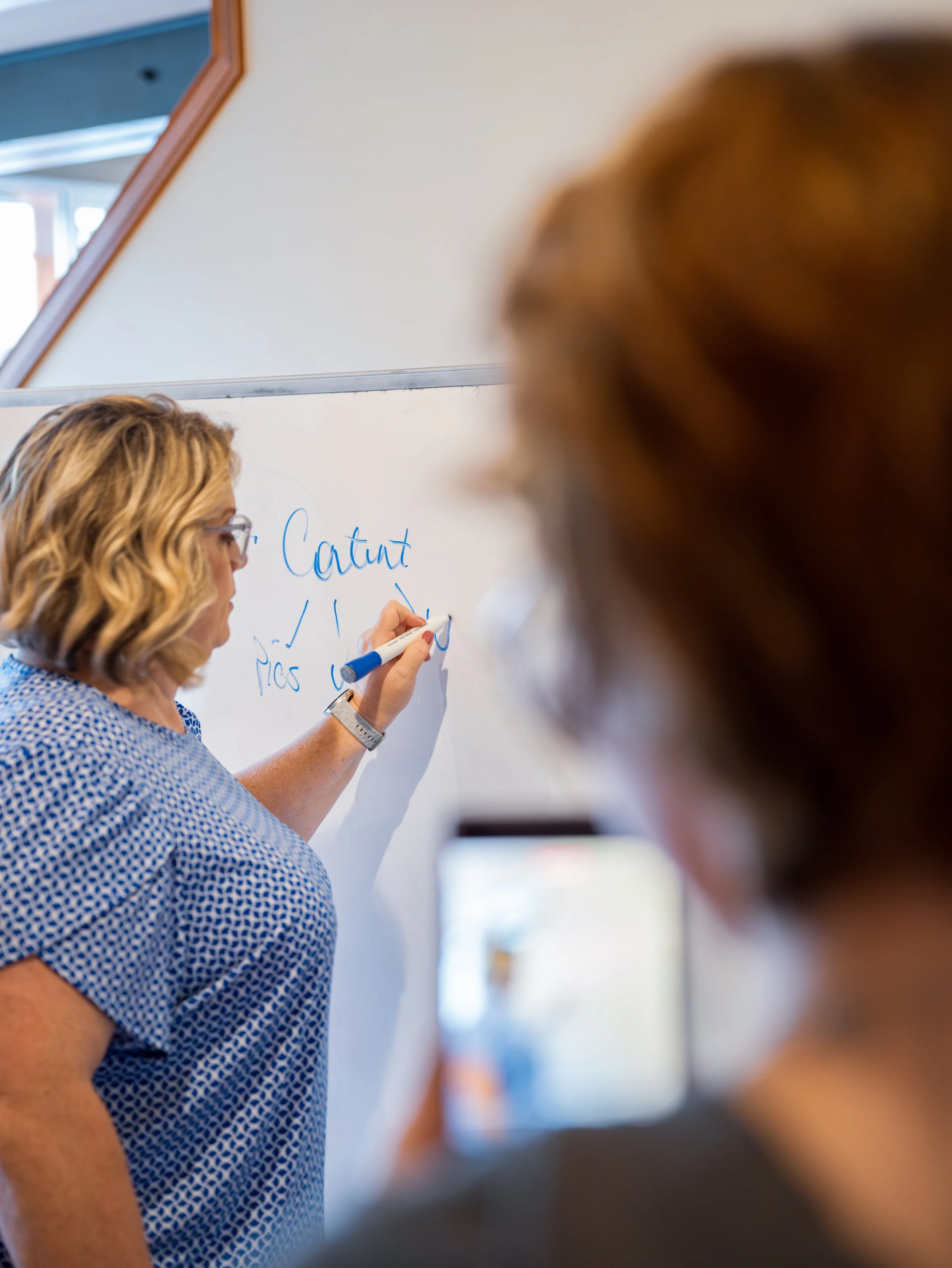 A woman with curly blonde hair and glasses writing on a whiteboard in a classroom or office. There is a partial view of another person in the foreground and a mirror on the wall reflecting the room.