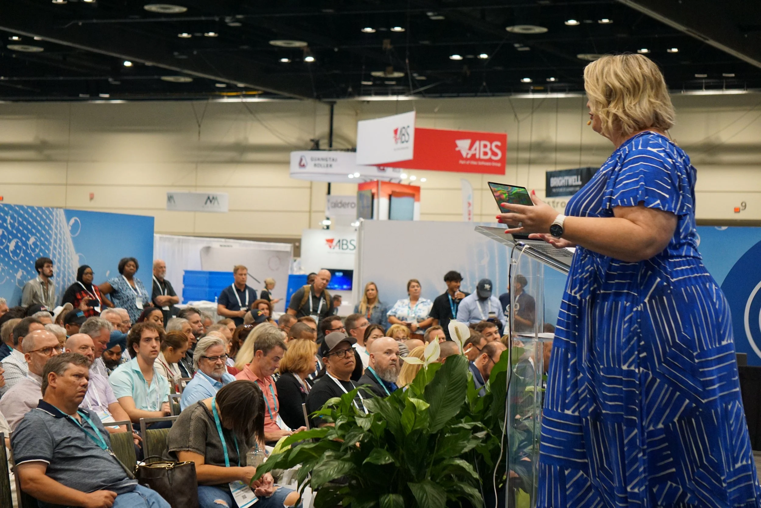 A woman in a blue striped dress speaking at a podium during a conference or event, with a large audience seated in front of her. There are banners and signs with 'ABS' and other logos in the background.
