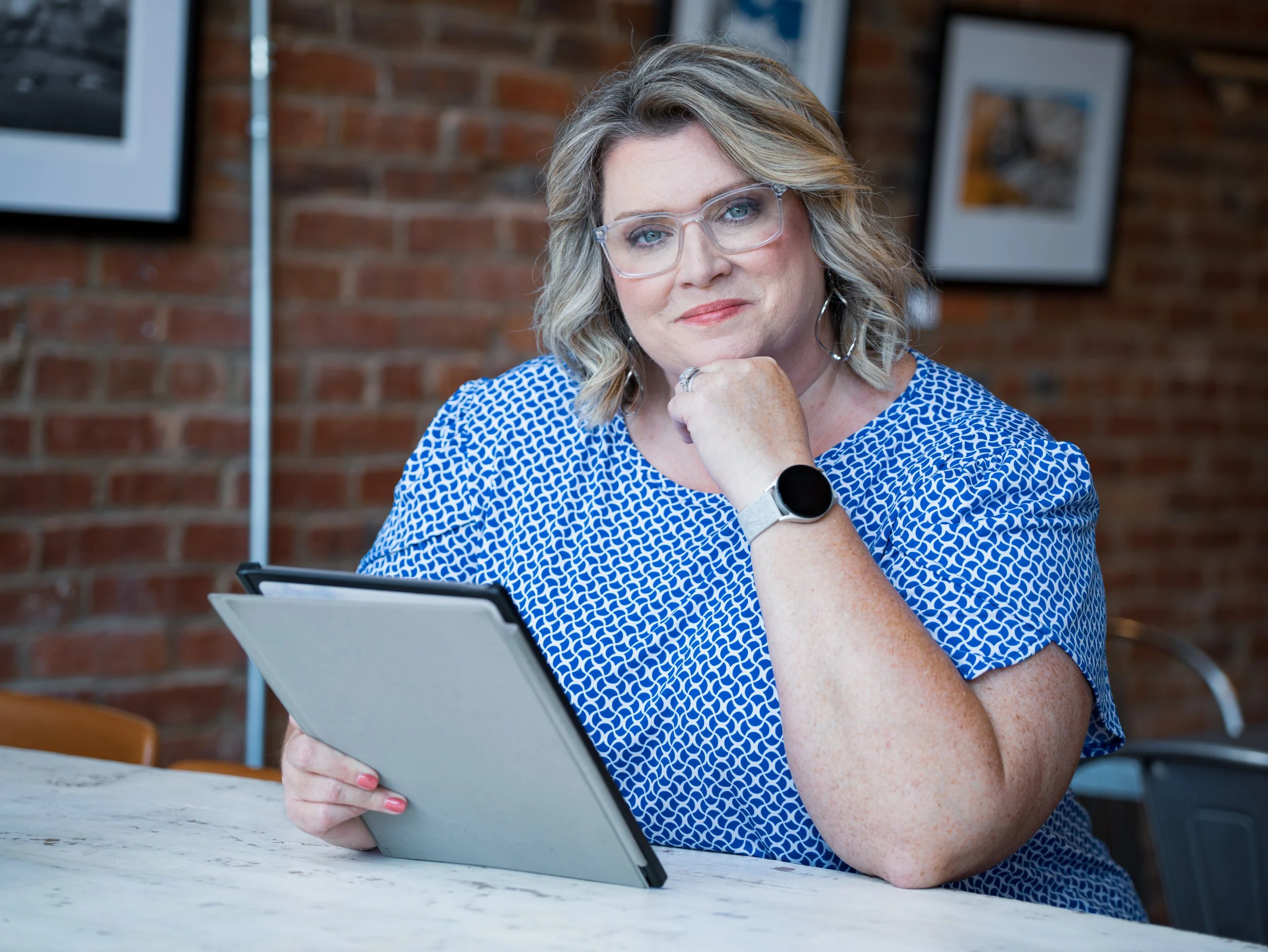 A woman wearing glasses and a blue patterned shirt is sitting at a table, holding a tablet with a brick wall and pictures in the background.