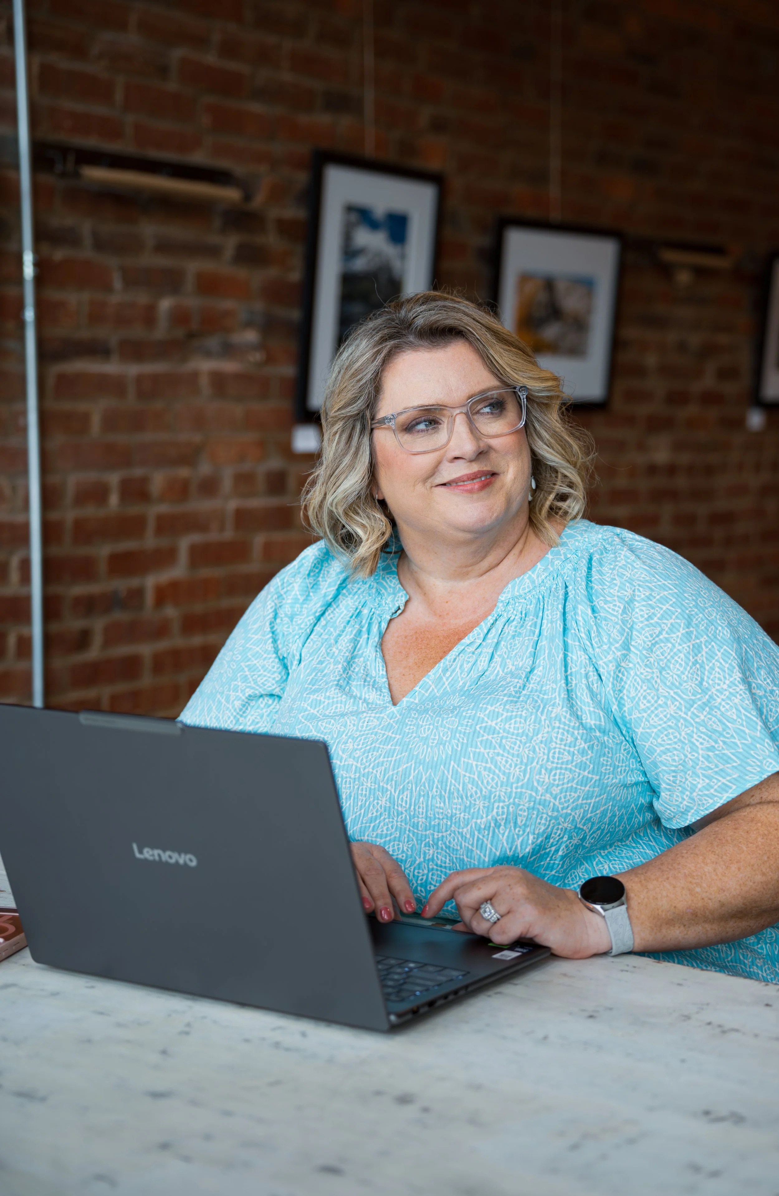 A woman with blonde, wavy hair and glasses, wearing a light blue patterned dress, sitting at a marble table in front of a Lenovo laptop, smiling and looking away. The background has a brick wall with framed pictures.