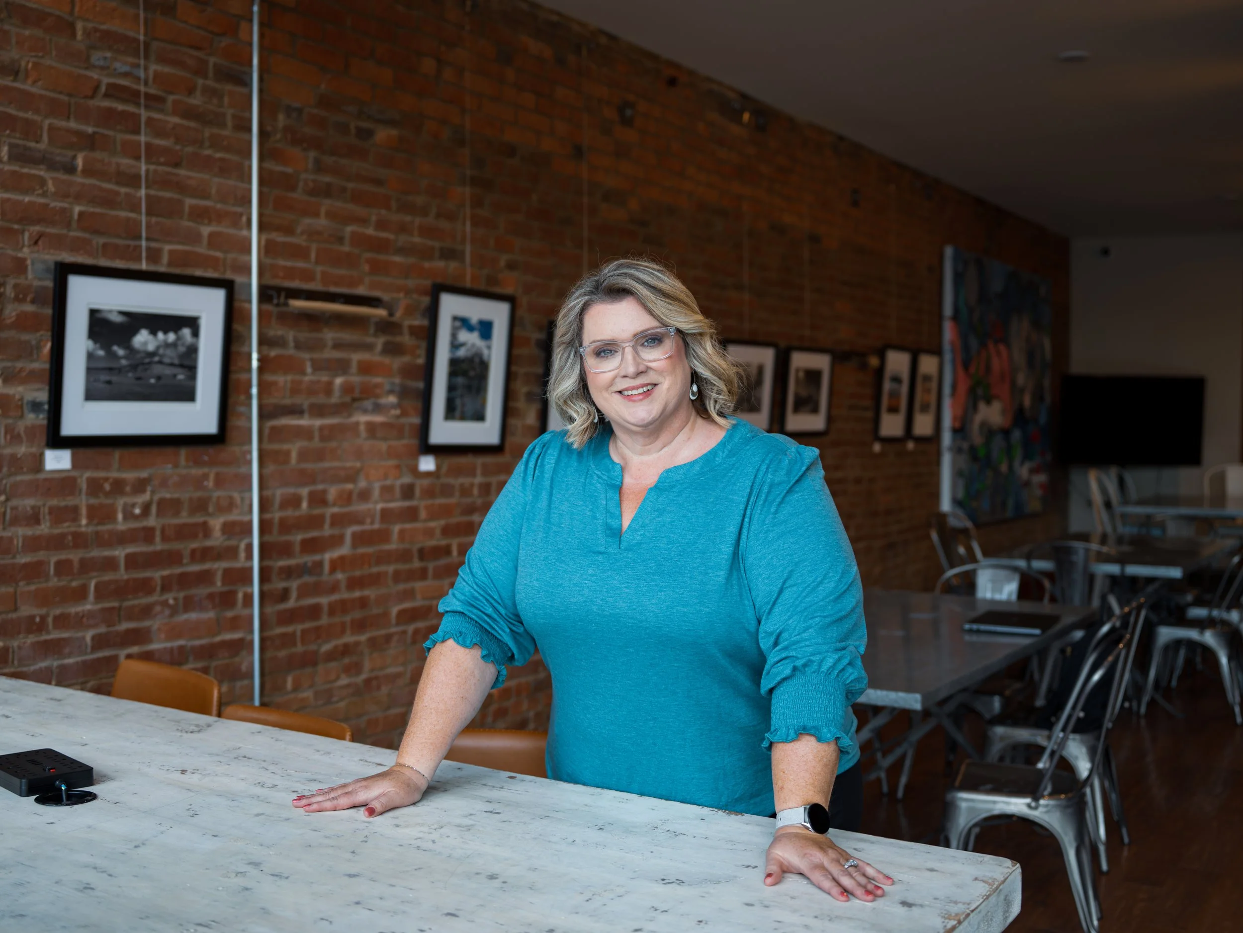 A woman with glasses and wavy blond hair wearing a turquoise top, smiling, leaning on a marble countertop in an art gallery or studio with framed photographs on a brick wall in the background.