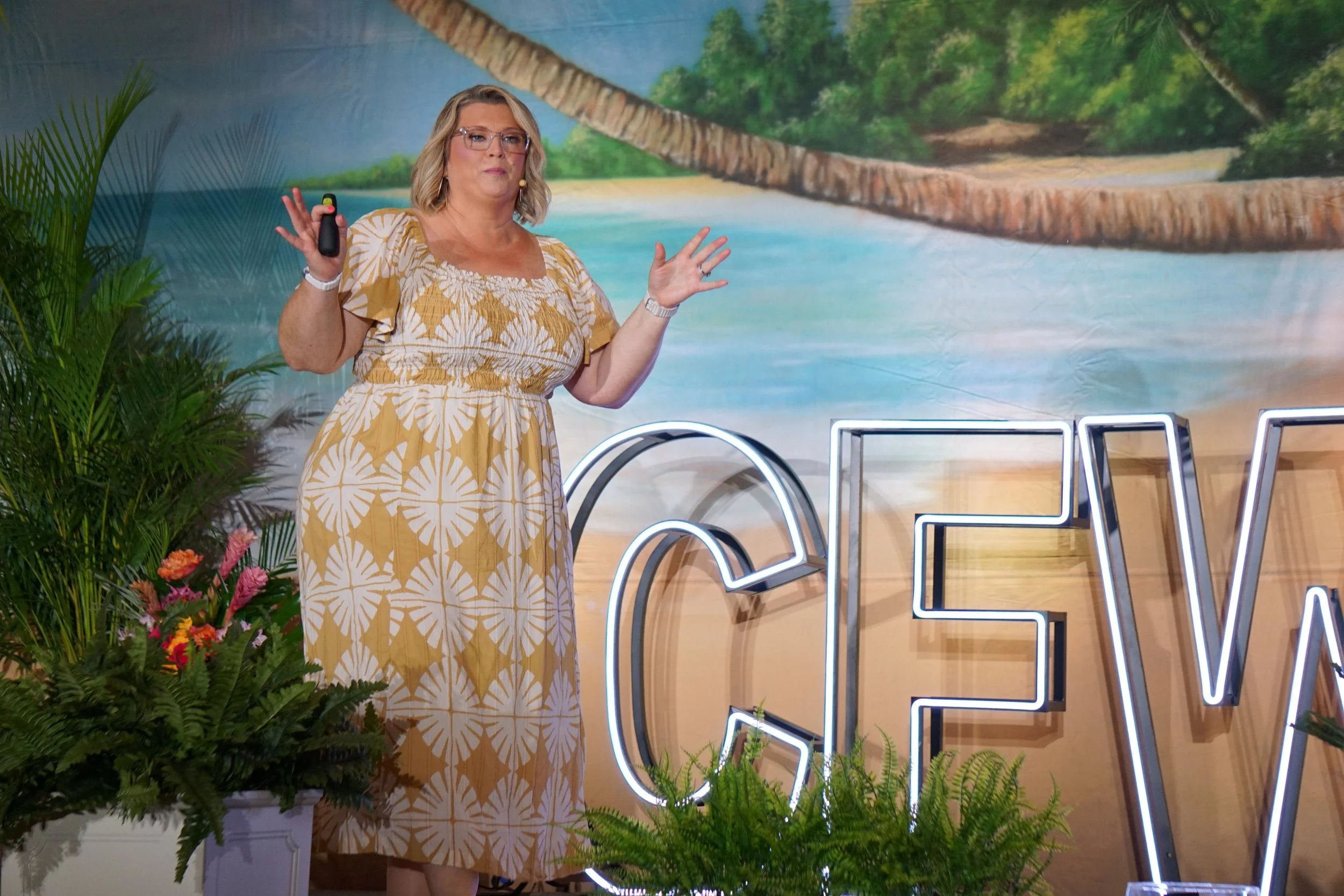 Woman with blonde hair and glasses giving a presentation in front of a tropical beach mural, surrounded by lush green plants and neon signs.
