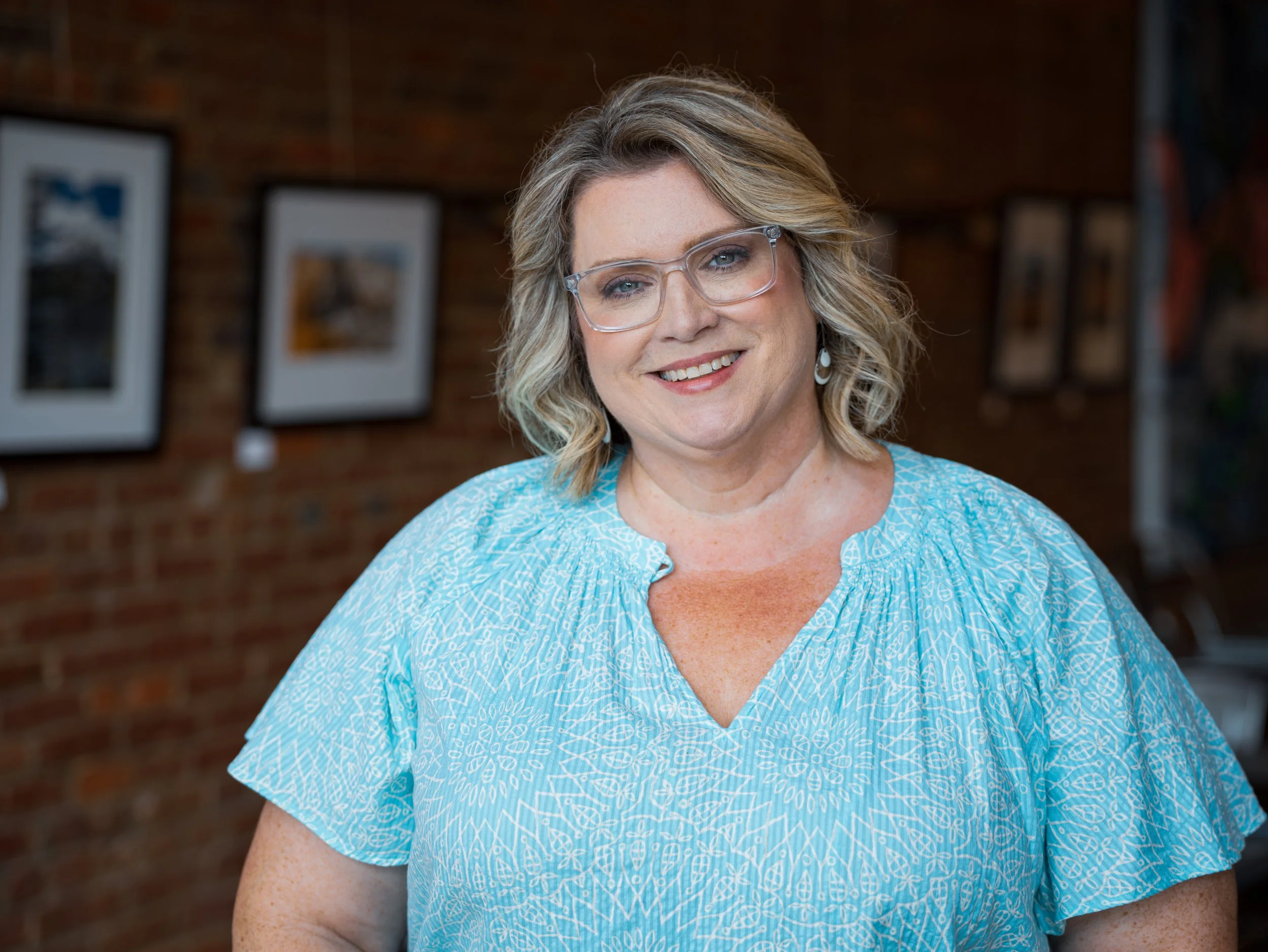 Smiling middle-aged woman with blonde wavy hair and glasses wearing a blue patterned dress, standing in front of a brick wall with framed photographs.